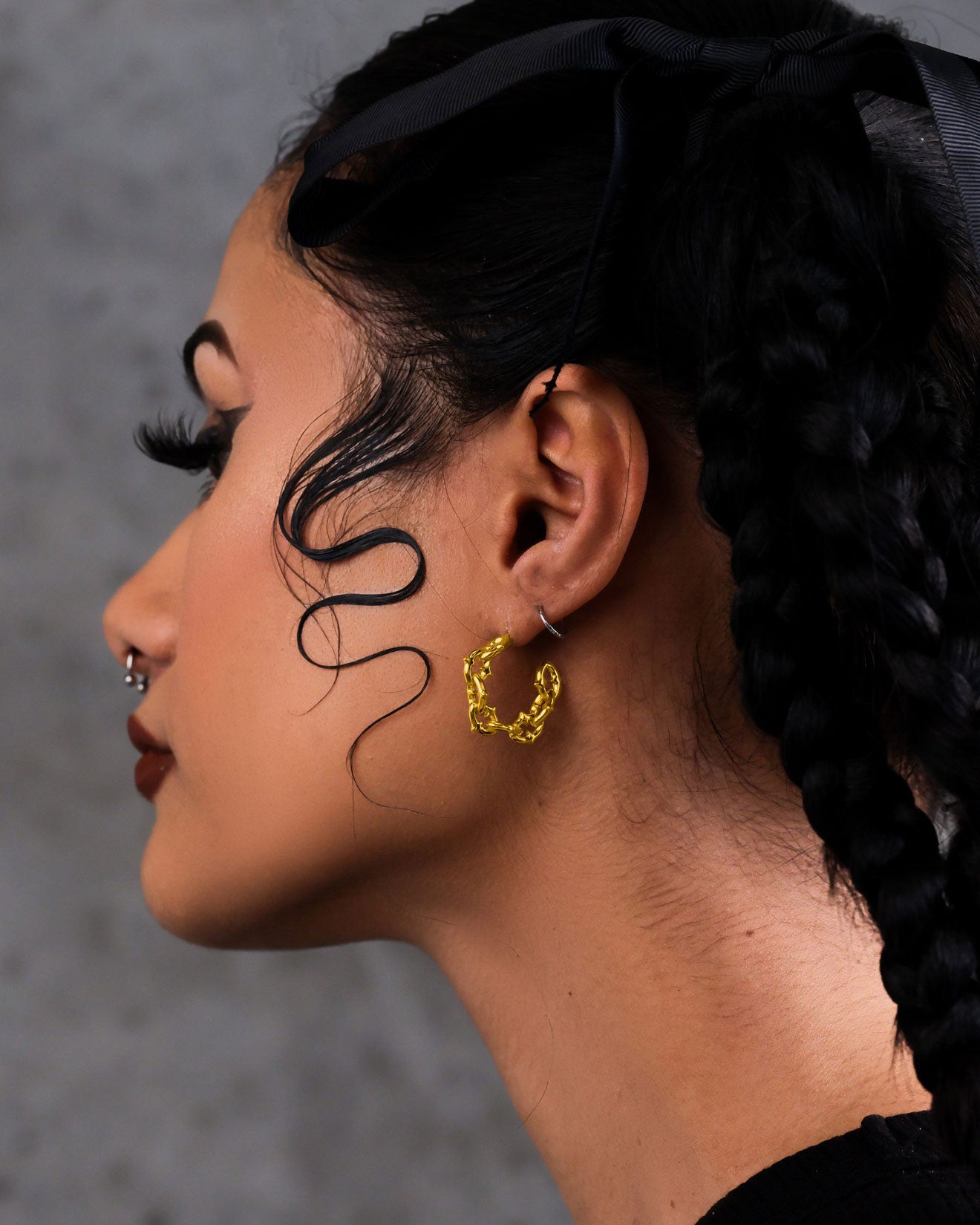 A person with braided hair, a black ribbon, and a face-framing curl wears "The Cathedral" Hoop Earrings (Gold) and a nose ring, shown in profile against a gray background.
