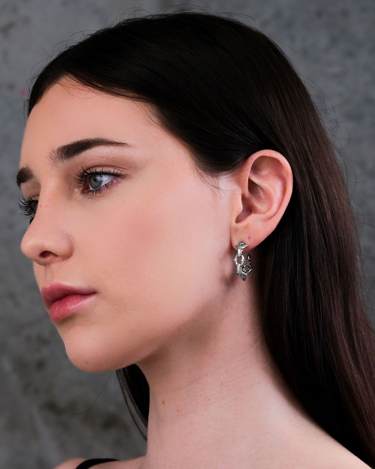 A woman with long brown hair is shown in profile against a gray textured background, wearing "The Cathedral" Hoop Earrings and a thin black strap top. Her makeup is natural and subtle.