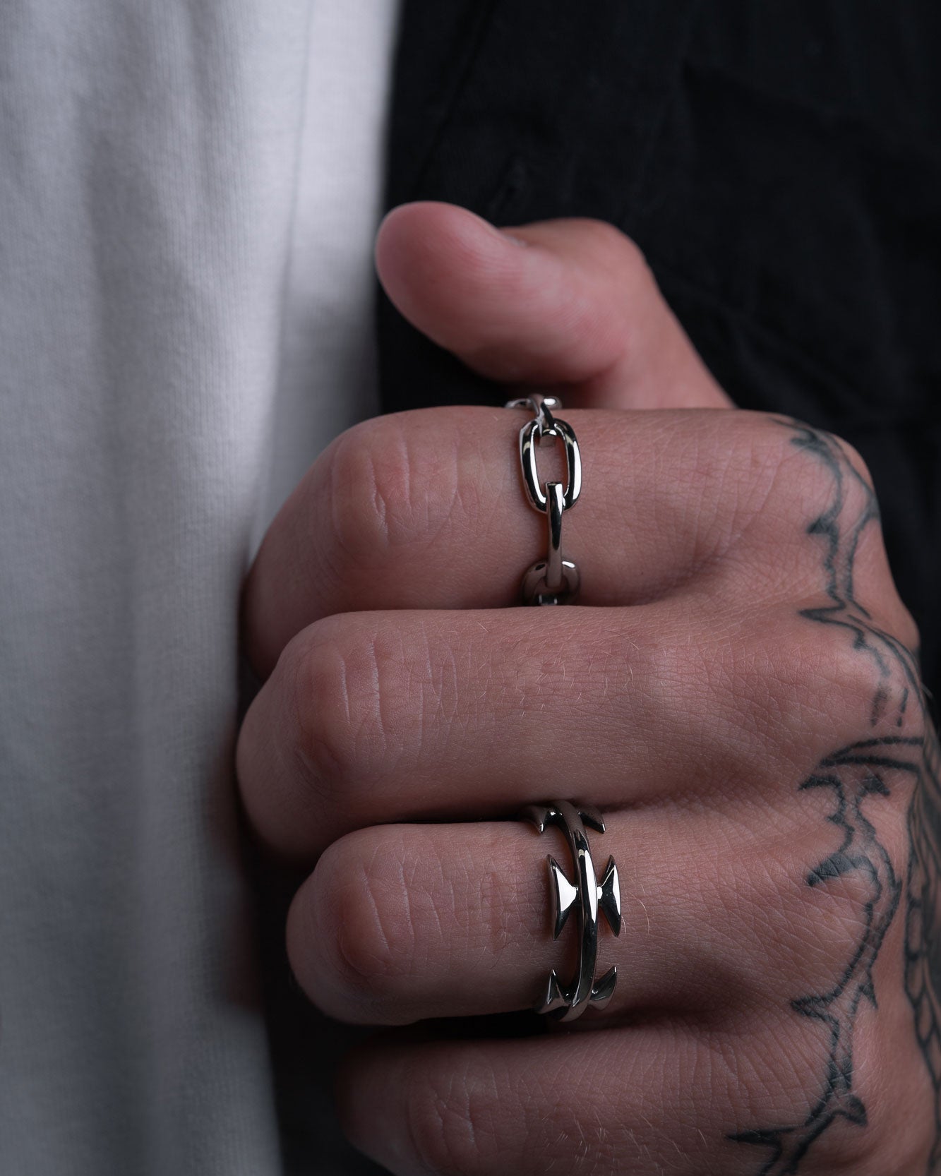 A tattooed hand wearing the Chained Ring—a chunky stacking style—along with a bold silver band, holds a silver chain. The person is dressed in a white shirt and black jacket.