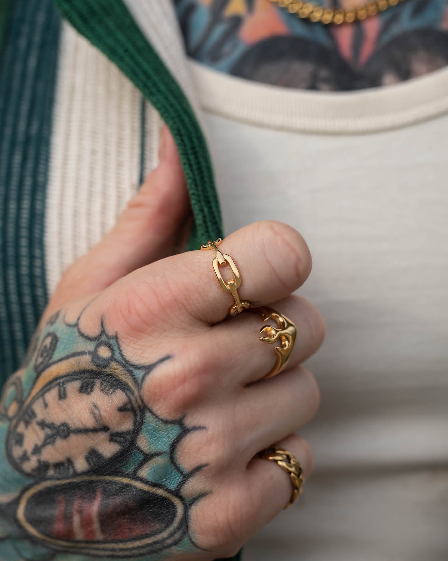 A tattooed hand adorned with the Chained Ring (Gold) and other gold rings holds green and white textured fabric, showcasing detailed pocket watch tattoos against a tattooed chest with a gold chain necklace.