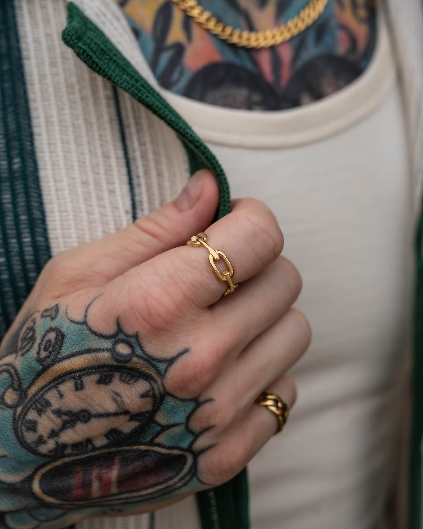 A close-up of a tattooed hand wearing two gold rings, including the Chained Ring (Gold), and holding the green collar of a zip-up jacket. A gold necklace and a pocket watch tattoo are also visible.