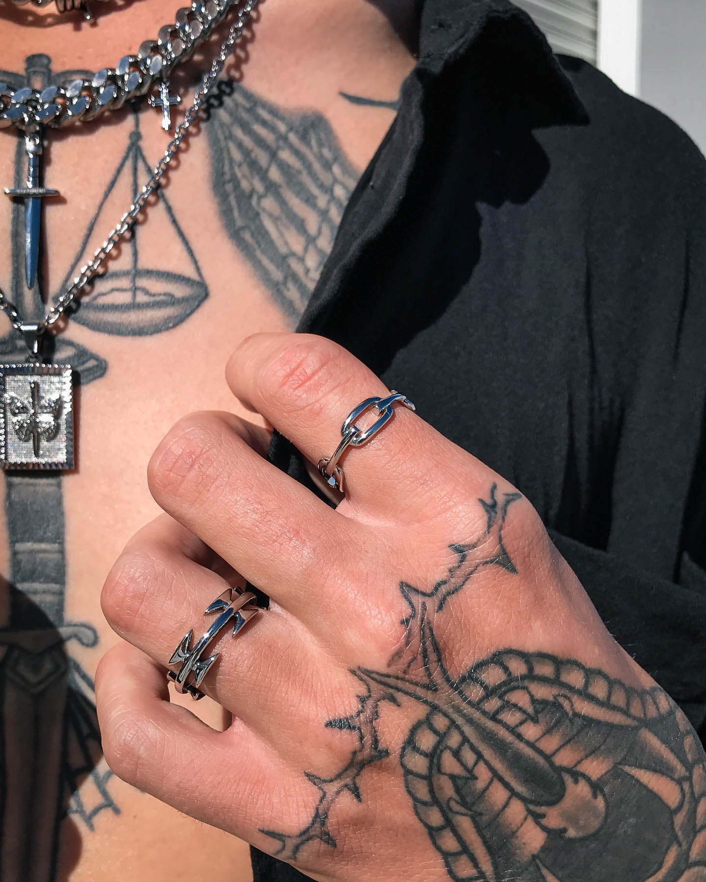 A person with bold black tattoos, wearing a Chained Ring and layered chain necklaces with a sword pendant, dressed in a partially open black shirt.