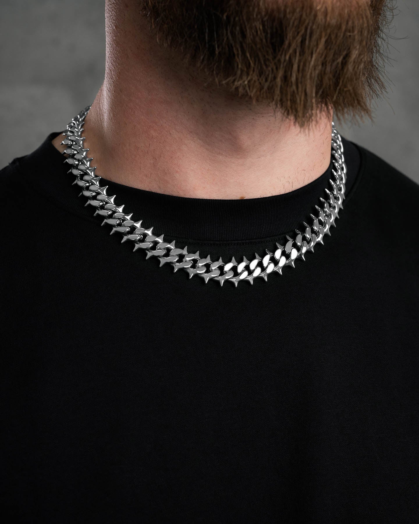 A bearded person wears the "Chainsaw" Spiked Cuban Chain necklace over a plain black shirt, with a blurred gray background.