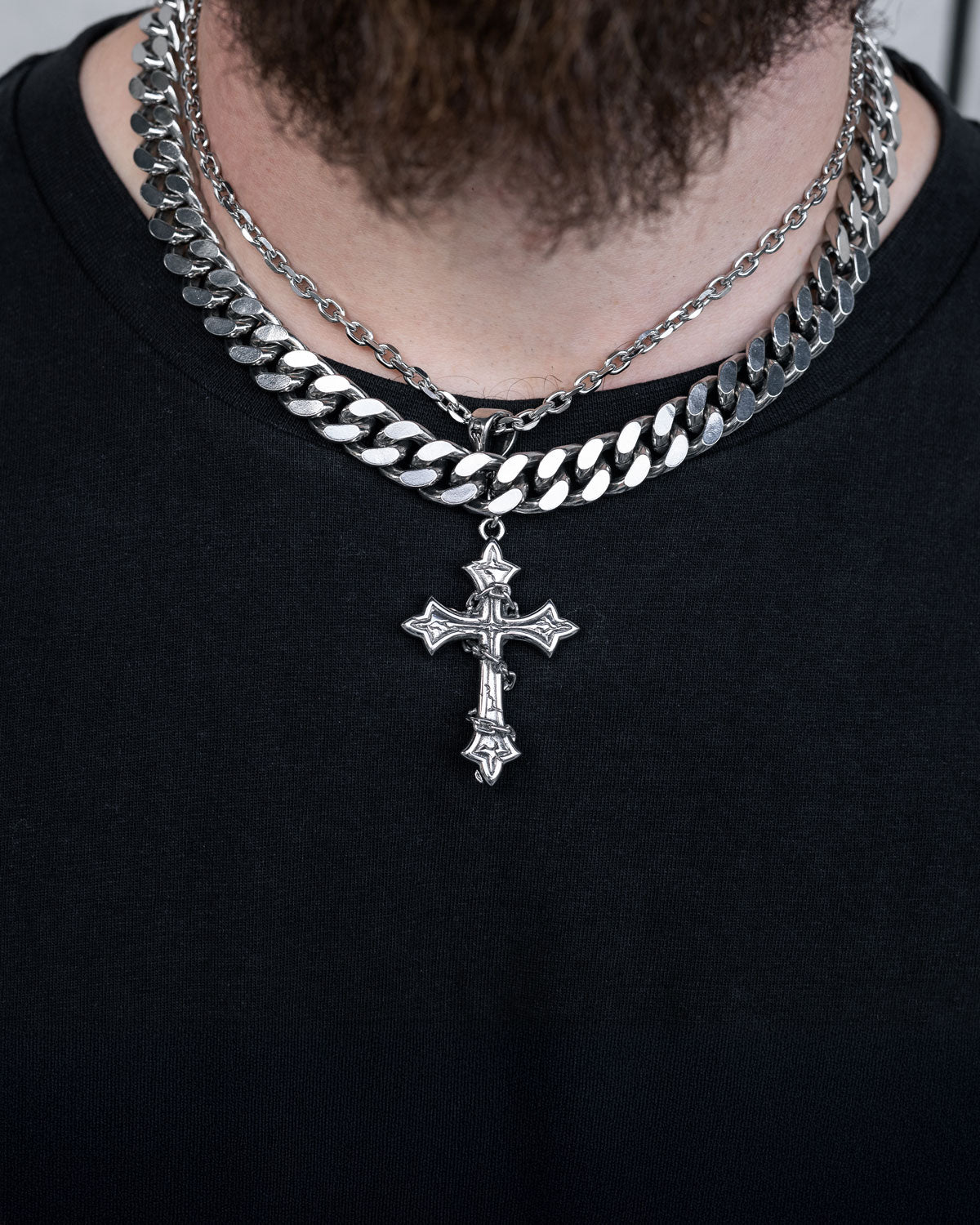A person wears a black shirt and the Chunky Ruins Set by Statement Collective, featuring two silver chains with a stainless steel cross pendant. Their beard is partially visible at the top of the image.