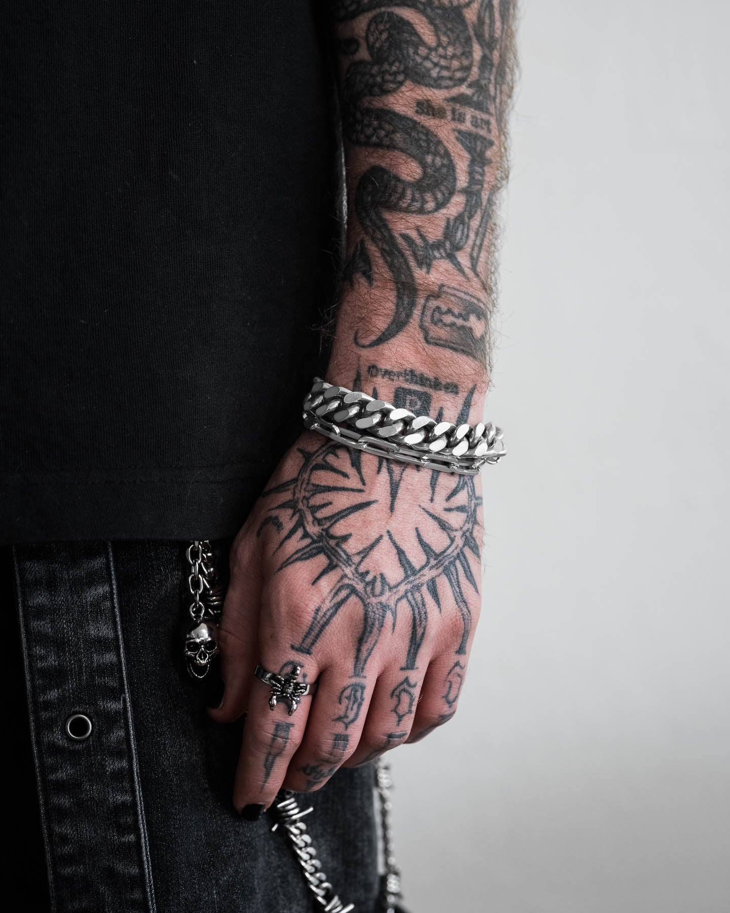Wearing black clothes with detailed arm and hand tattoos, this person sports a 13mm Cuban Cluster Bracelet, bold lock chain accessories on their dark jeans, a skull ring, and has black-painted fingernails.