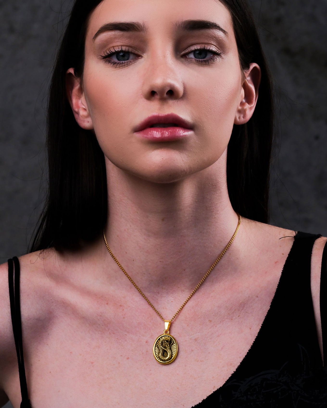 A woman with straight dark hair wears a black top and the Snake Charmer Pendant (Gold), facing forward with a neutral expression against a dark background.