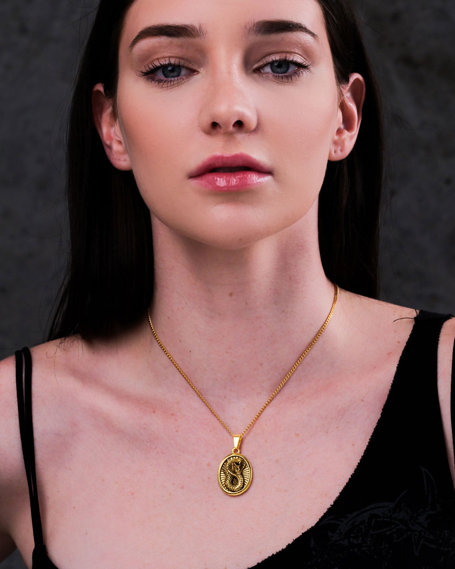 A woman with straight dark hair, dressed in black, wears the Snake Charmer Pendant (Gold) and looks directly at the camera with a neutral expression against a dark background.
