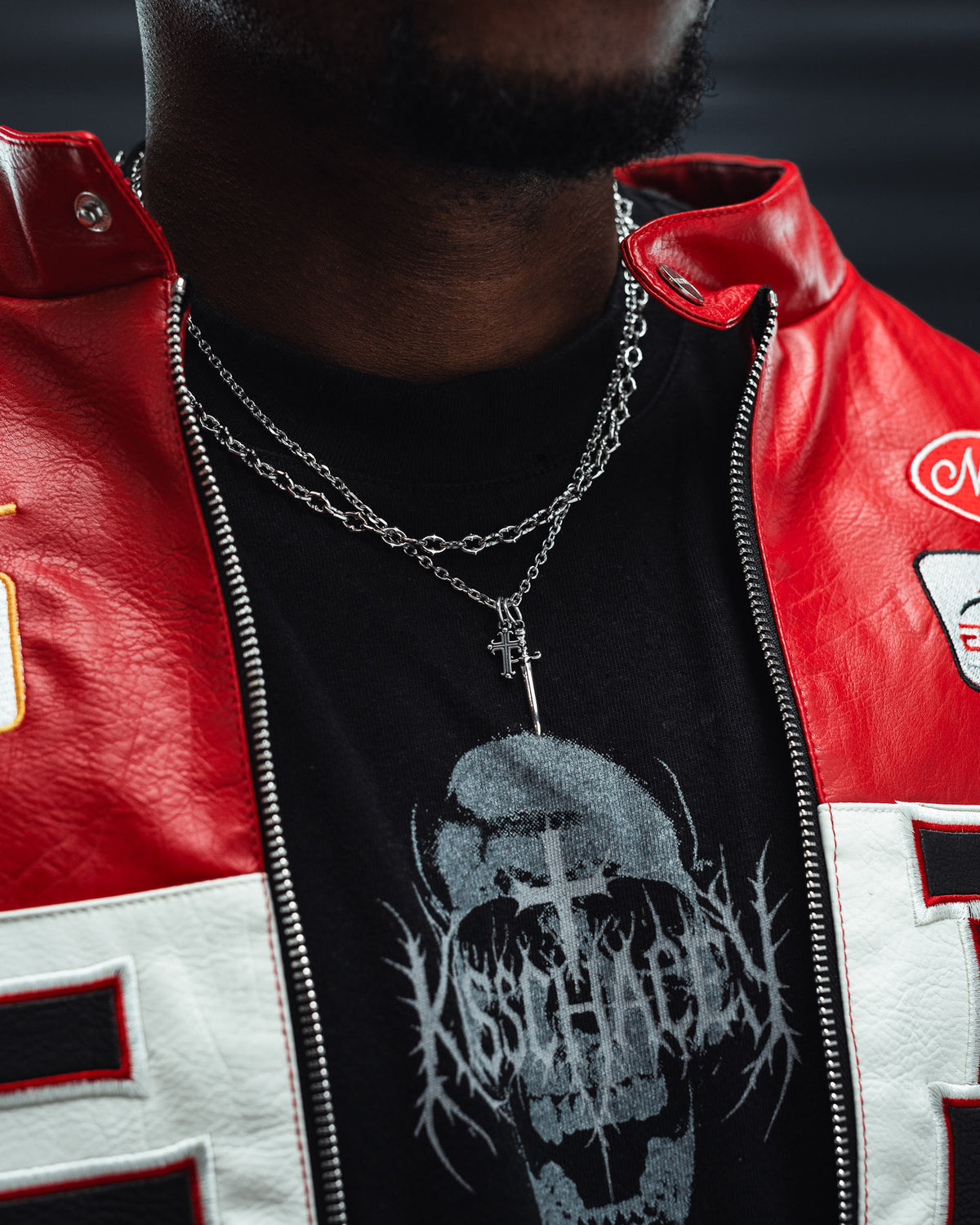 Close-up of a person wearing a red and white jacket, black graphic t-shirt with a skull design, and the Statement Collective Cross & Dagger Necklace layered for a dark romance jewelry style. The person’s face is not visible, emphasizing the bold statement piece.