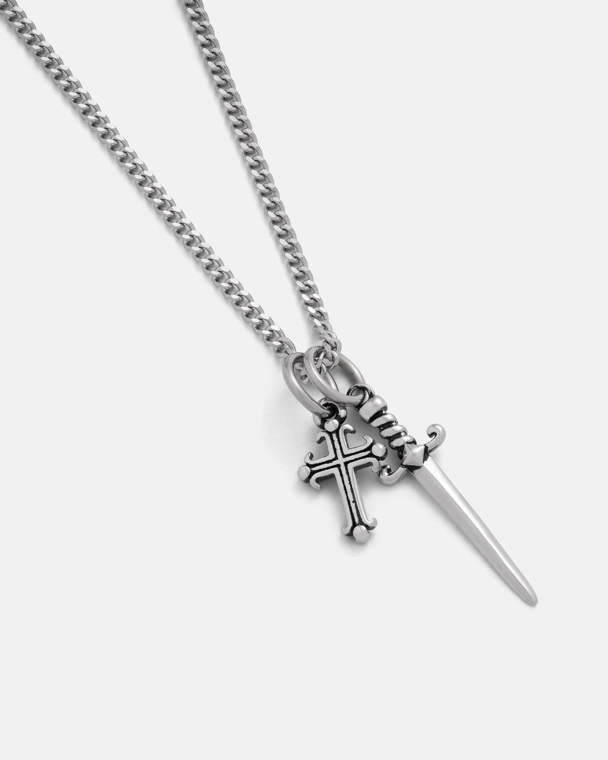 The Cross & Dagger Necklace by Statement Collective features a silver stainless steel chain adorned with two pendants—a decorative cross and a finely detailed dagger—hanging side by side against a plain light background. Ideal for those captivated by dark romance aesthetics, this piece seamlessly blends elegance with bold, edgy appeal.