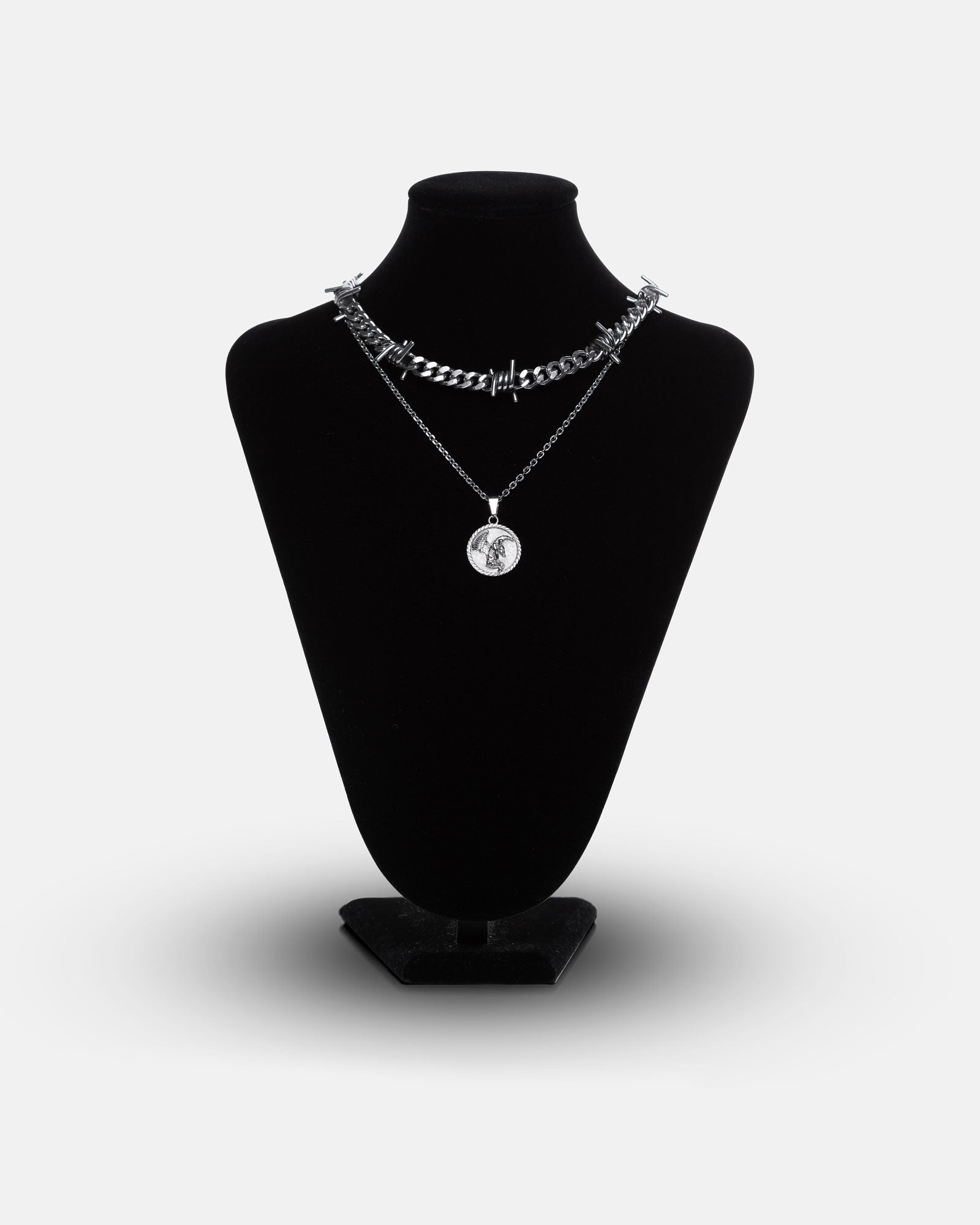 A black necklace display stand showcases the Crucifix Set: one chunky barbed wire necklace and one slim chain with a circular fallen angel medallion pendant.