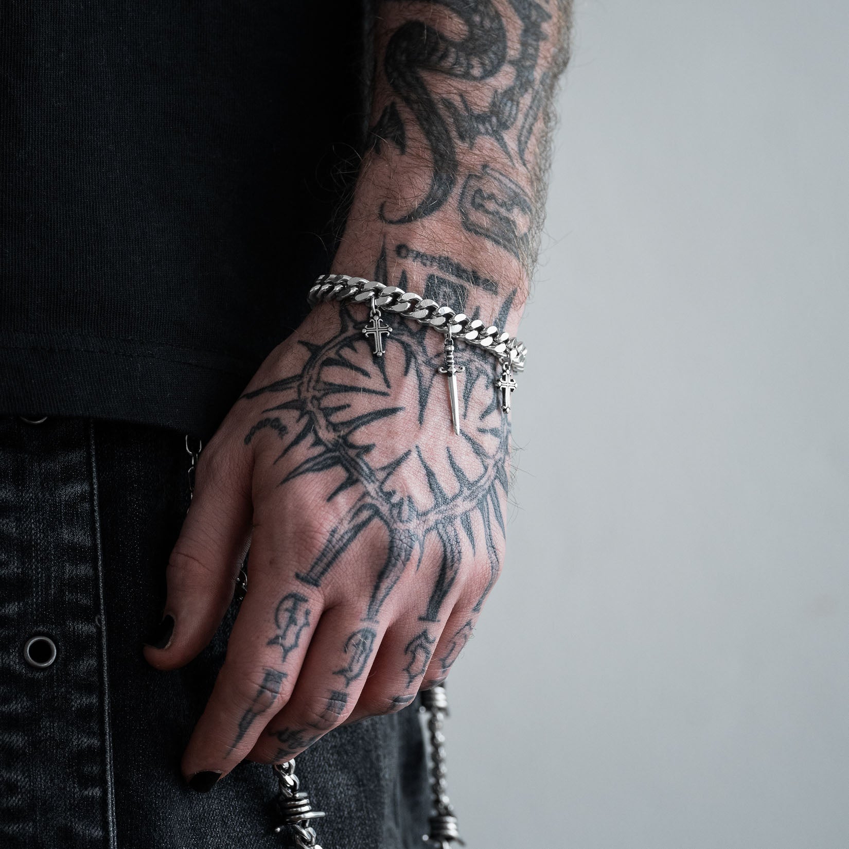 Hand with tattoos wearing a silver chain bracelet with dagger and cross charms on a plain background