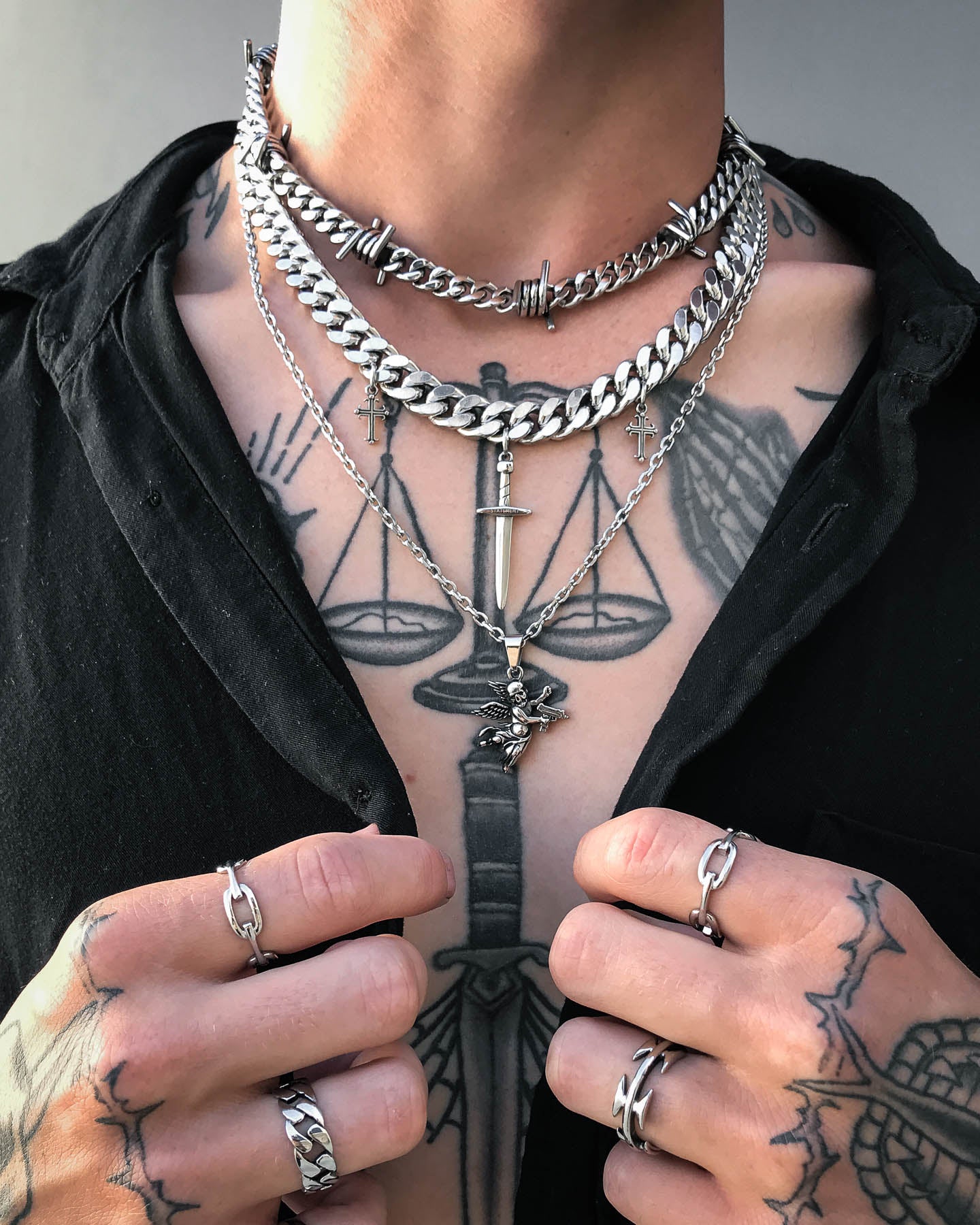 A person wearing multiple chunky silver chain necklaces, including the "Crusader" 11mm Cuban Chain and a punk-style necklace with cross and dagger pendants, plus silver rings and a black open-chest shirt that reveals a scales of justice tattoo.