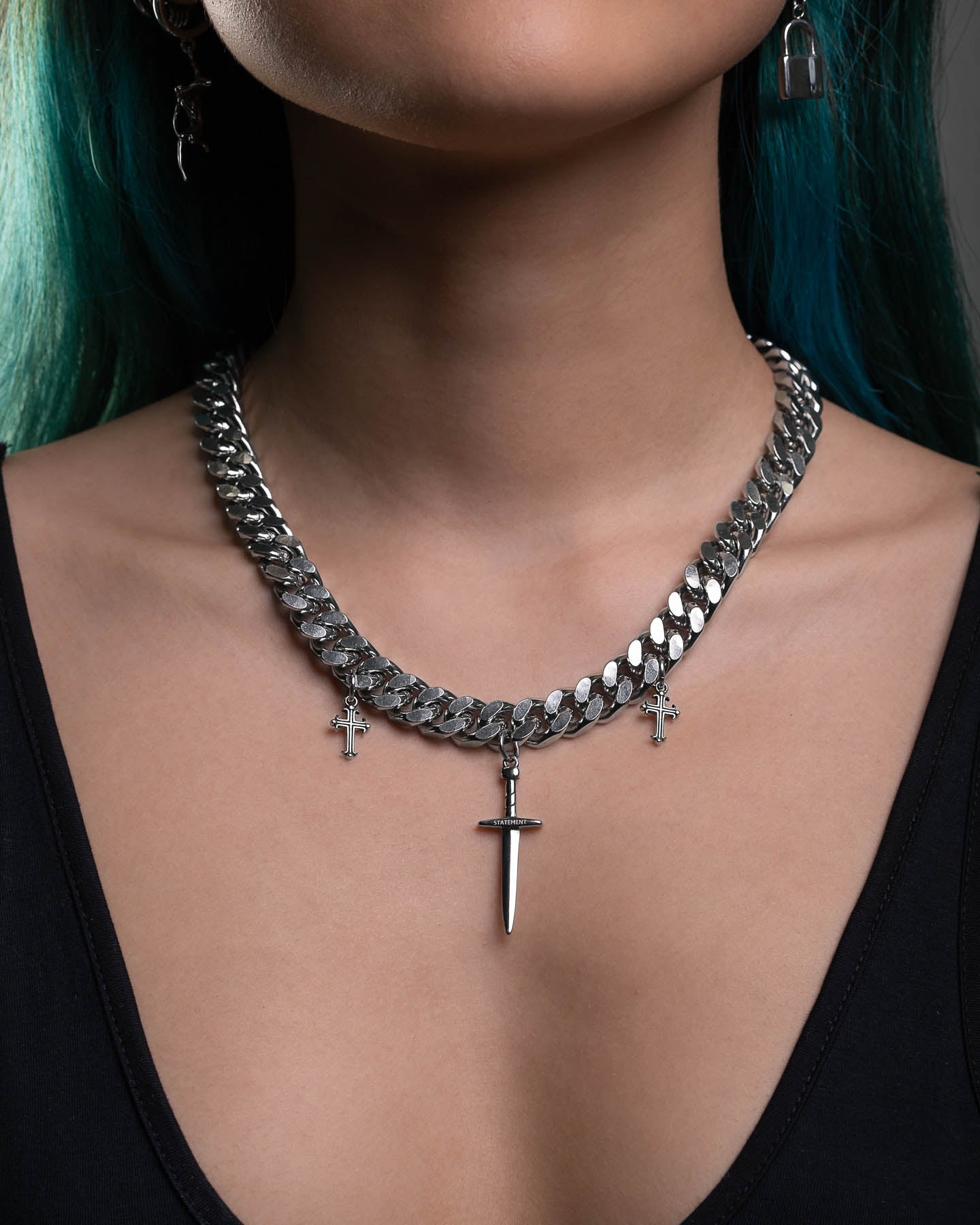 Wearing the "Crusader" 11mm Cuban Chain with three dagger pendants, paired with a black top, blue-green hair, and visible earrings highlighting their neckline for a bold punk-inspired look.