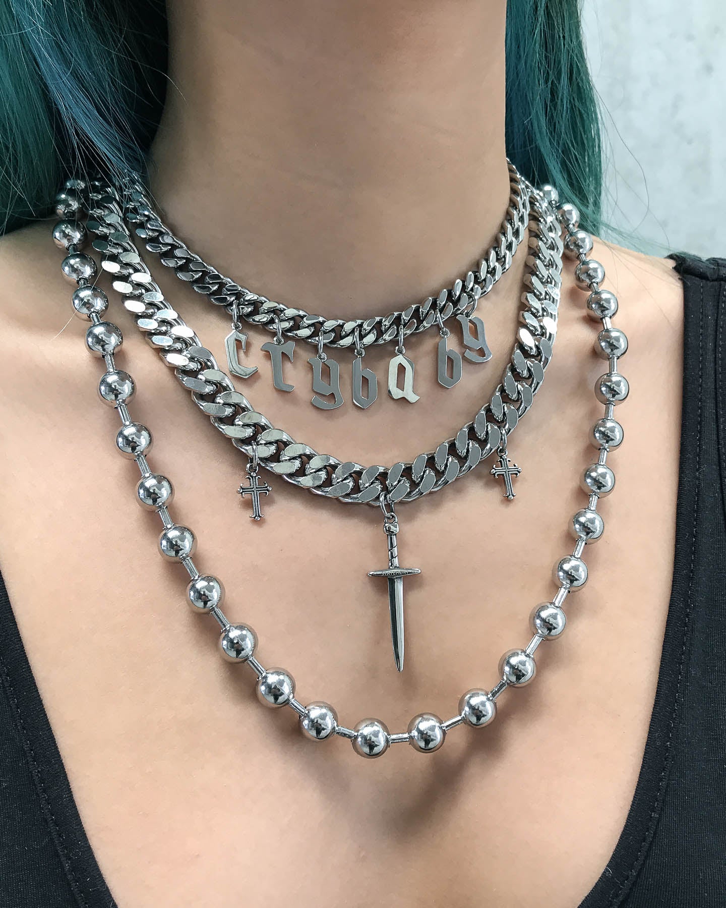 A person with blue-green hair and a black top wears festival jewelry: the Crybaby Stack—three layered silver necklaces including a beaded choker, a dagger pendant with cross charms, and a chunky gothic-lettered Crybaby necklace.