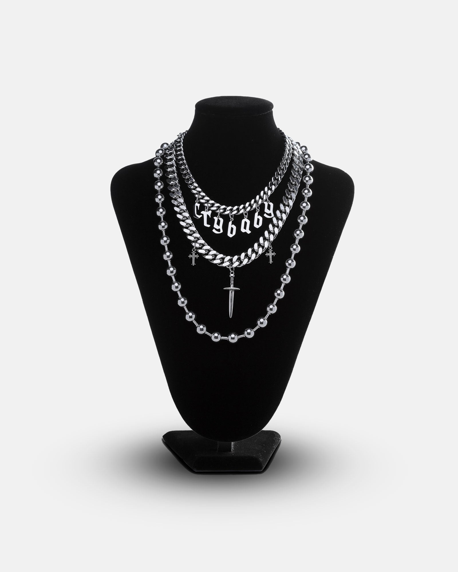 The Crybaby Stack features layered silver chains—a bold crybaby necklace, a dainty choker, and cross and dagger pendants—shown on a black velvet stand for the perfect festival jewelry look against a plain white background.