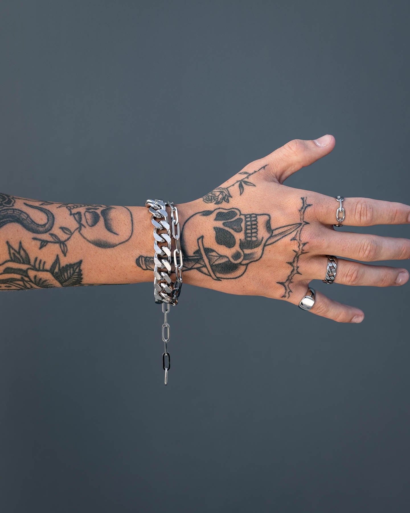 A tattooed hand wearing silver rings and the 13mm Cuban Cluster Bracelet with a box lock is shown against a plain gray background. The tattoos feature a skull, flowers, and patterns, with fingers slightly spread.