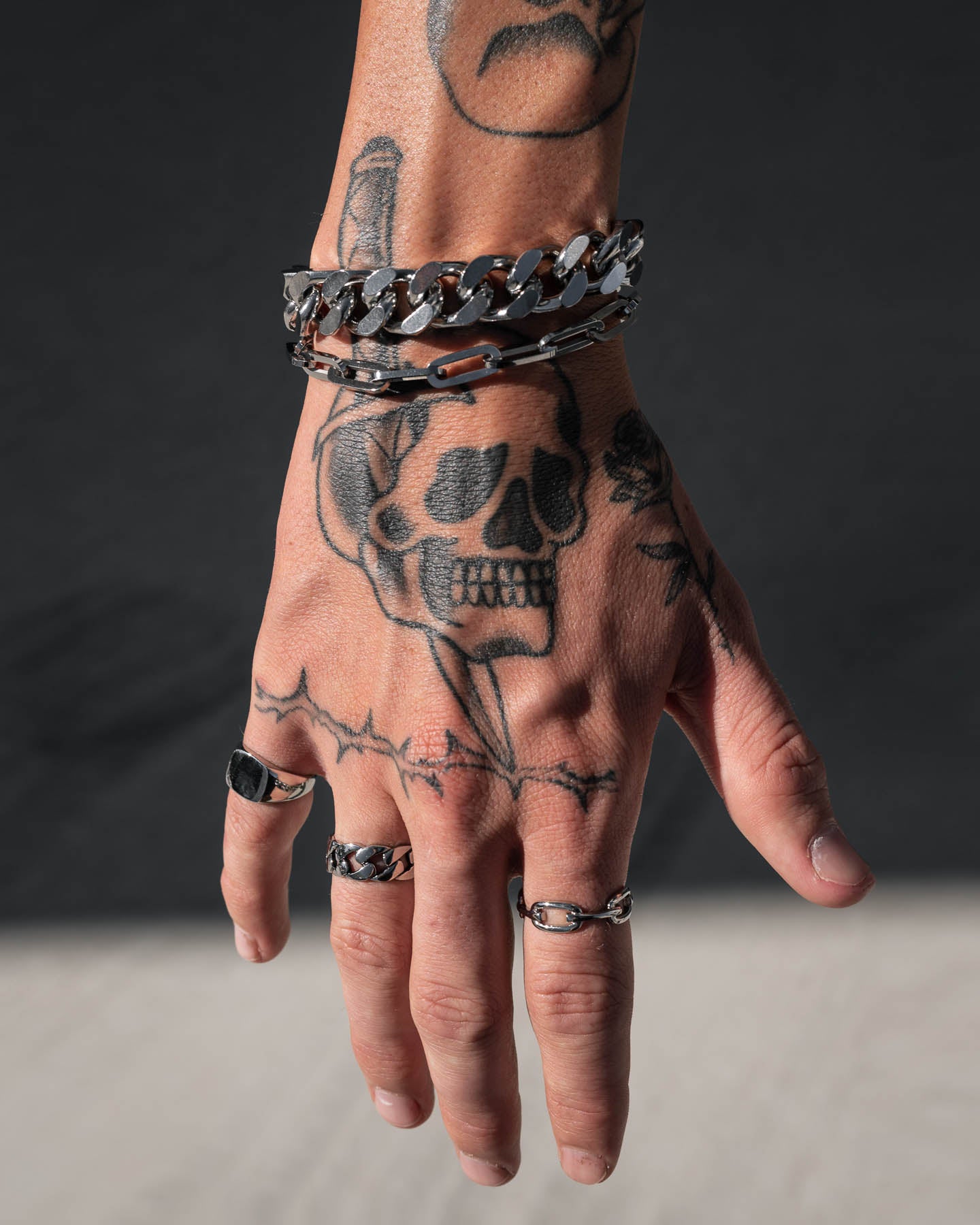 A hand with skull and barbed wire tattoos wears chunky silver rings and the bold 13mm Cuban Cluster Bracelet against a dark background.