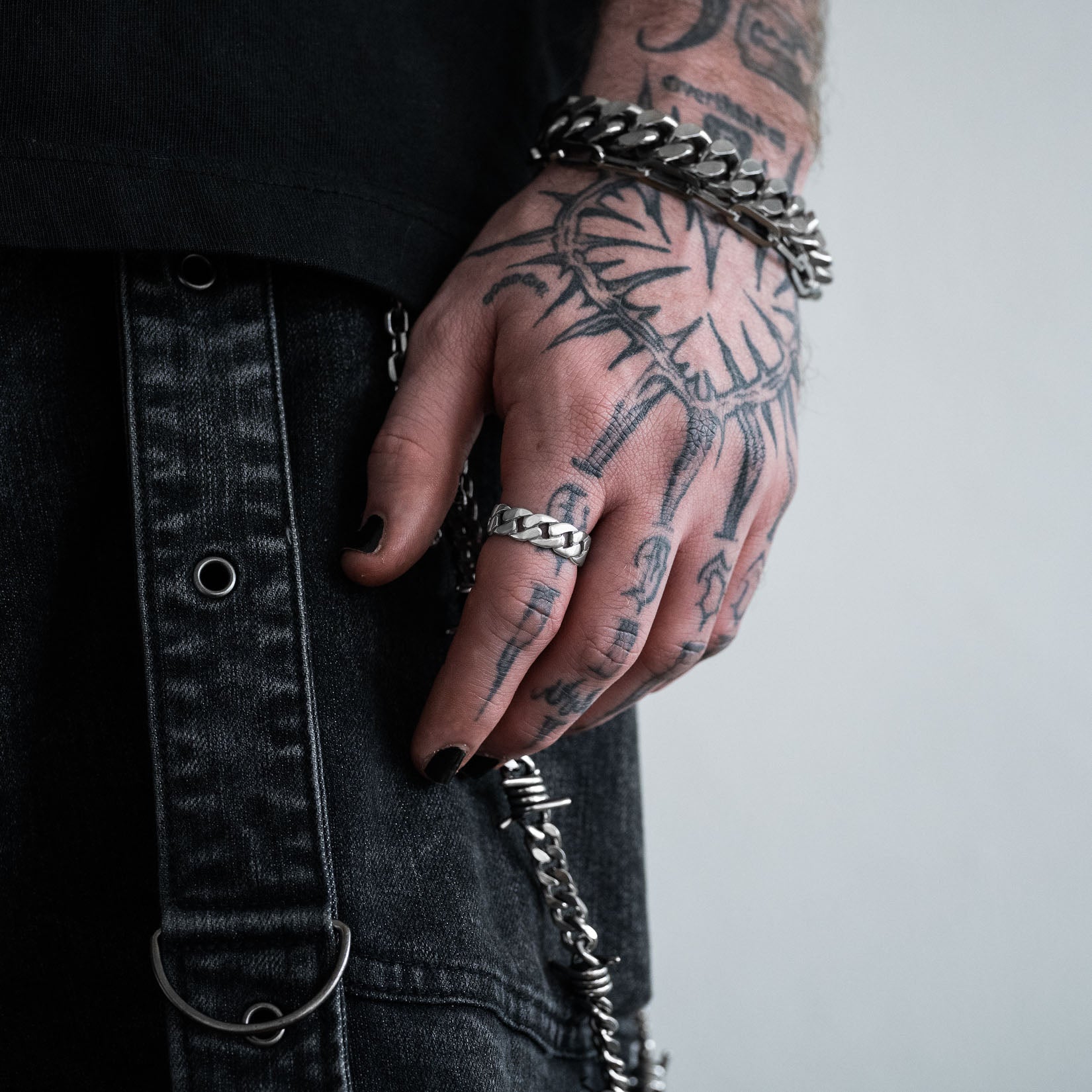 Hand with tattoos wearing a silver chain bracelet and cuban ring on a plain background by statement