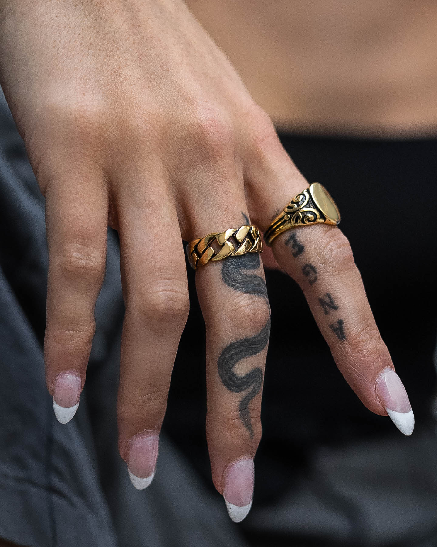 A hand with long nails, snake and letter tattoos, wears a chunky 18k gold ring and the Cuban Link Ring (Gold) on the middle and ring fingers, paired with a sleek black outfit.