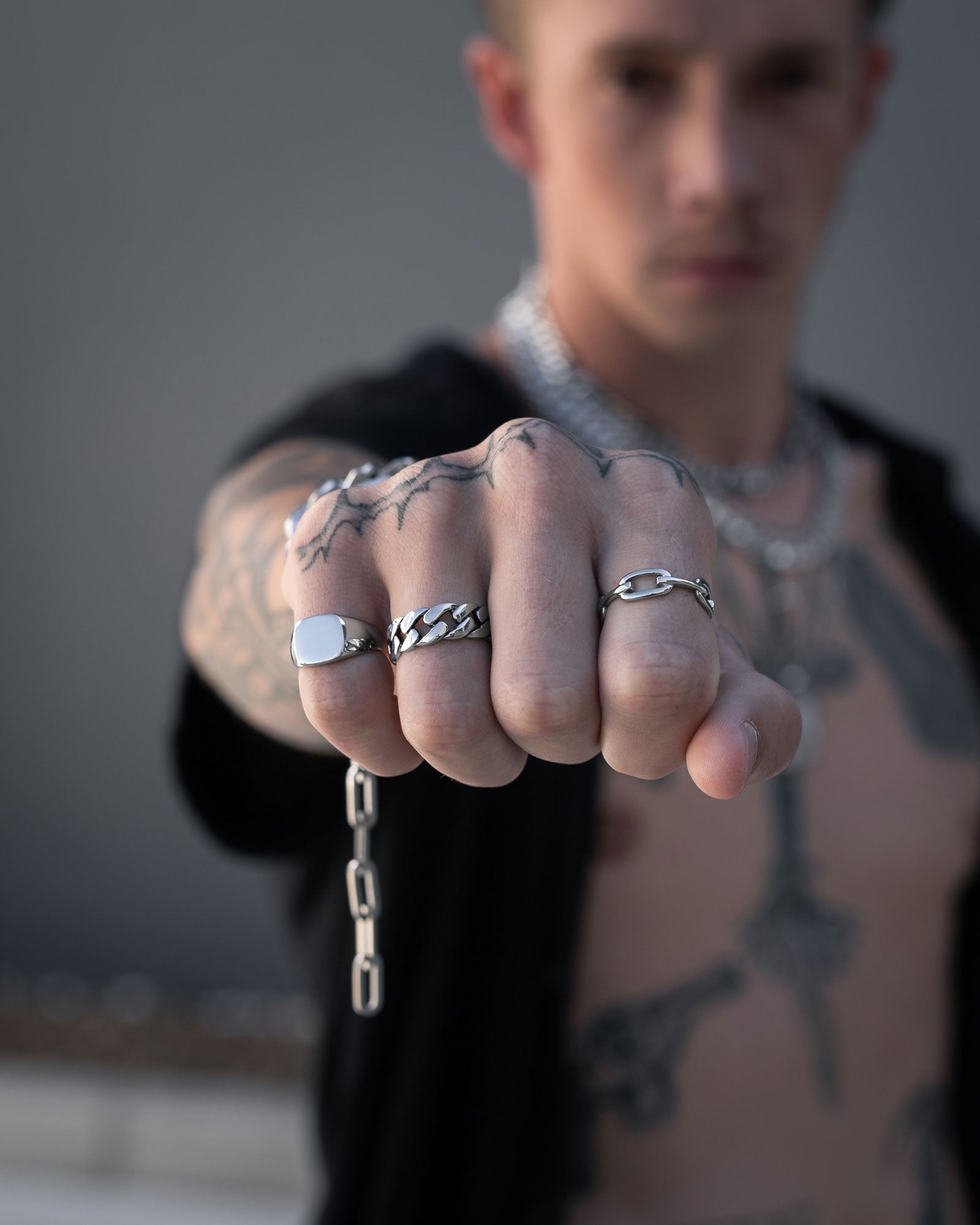 A tattooed person extends a closed fist toward the camera, displaying several silver rings—most notably the Cuban Link Ring—and a chain bracelet. The background is blurred and their face remains out of focus.