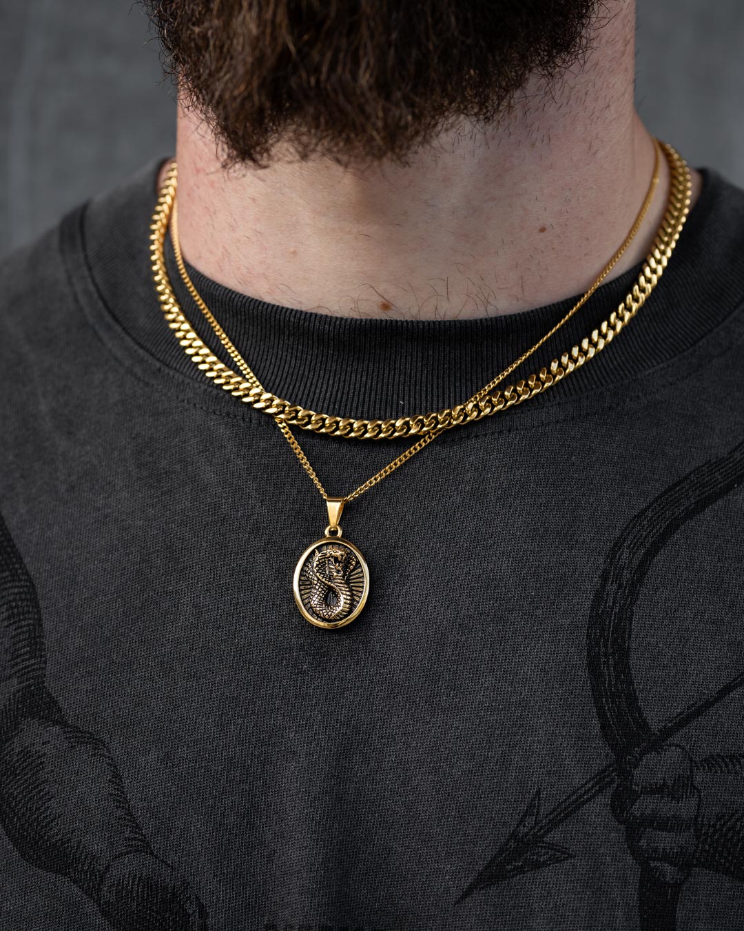 A close-up of a person wearing Statement Collective's Cuban Set (Custom Pendant Choice) in gold, featuring a round dragon pendant. They wear a dark shirt with a subtle graphic print and part of their beard is visible.