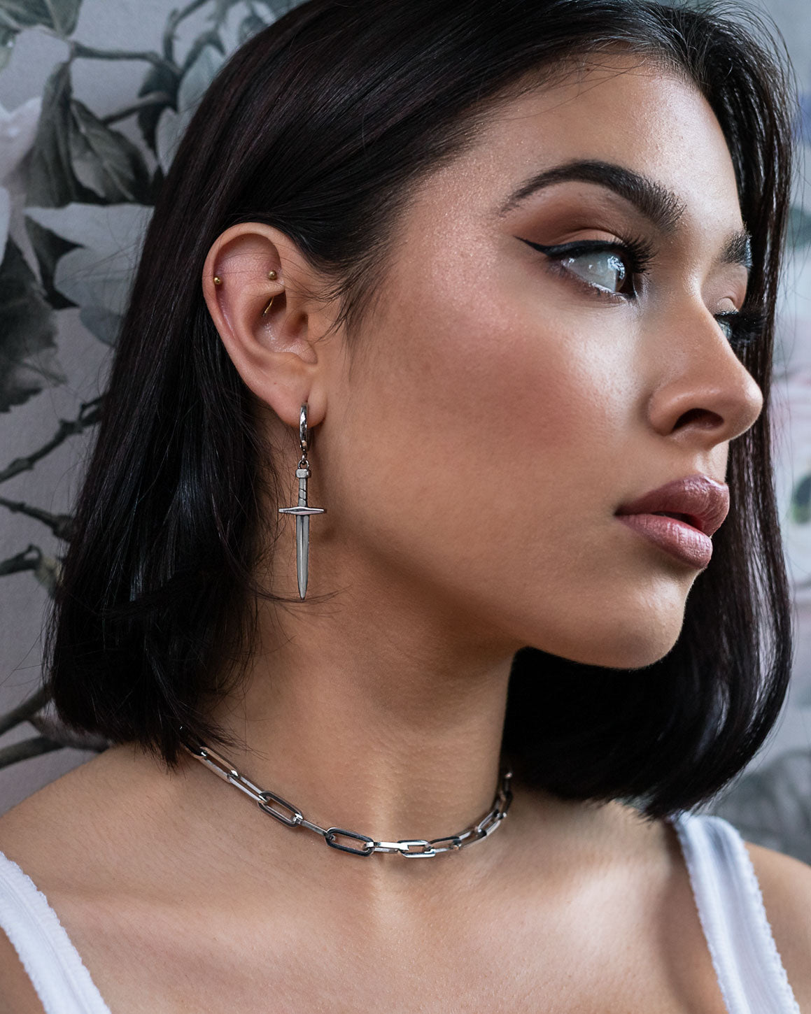 A woman with straight dark hair and winged eyeliner looks to the side. She wears a silver paperclip chain necklace, a white top, and the Dagger Earring—bold statement jewelry—set against floral wallpaper.