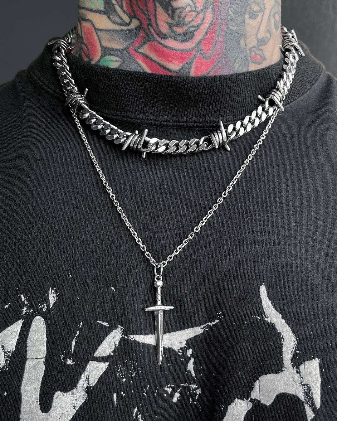 A person with colorful neck tattoos wears two silver necklaces: a chunky barbed chain choker and a fine chain featuring the Dagger Pendant. They are dressed in a black shirt with a distressed white graphic.