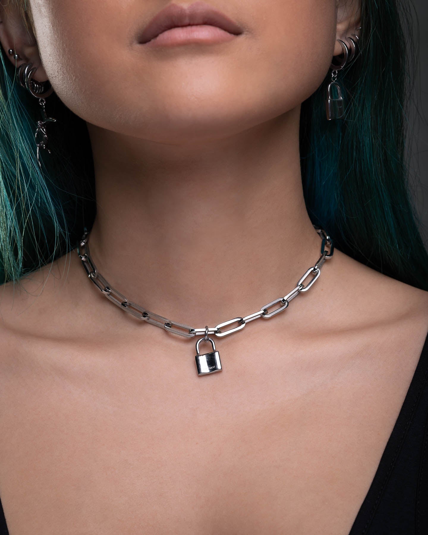 A person with teal hair wears The Dainty Charm Choker (Custom Pendant Choice) and matching padlock earrings, paired with a black top. The close-up highlights the jewelry and lower face.