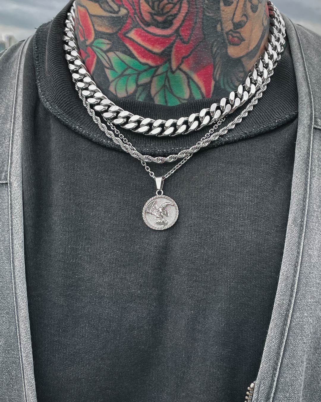 Wearing the Date Night Stack—layered silver chains, featuring a rope chain and coin pendant—a person with a colorful rose and leaf neck tattoo styles them over a black shirt and gray jacket.