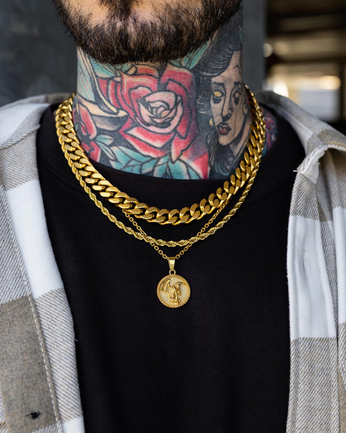A person wears Statement Collective’s Date Night Stack (Gold), a trio of gold chains with an 18k Gold Pendant Necklace, over a black shirt and plaid overshirt; colorful tattoos—a rose and woman’s face—adorn their neck and chest.