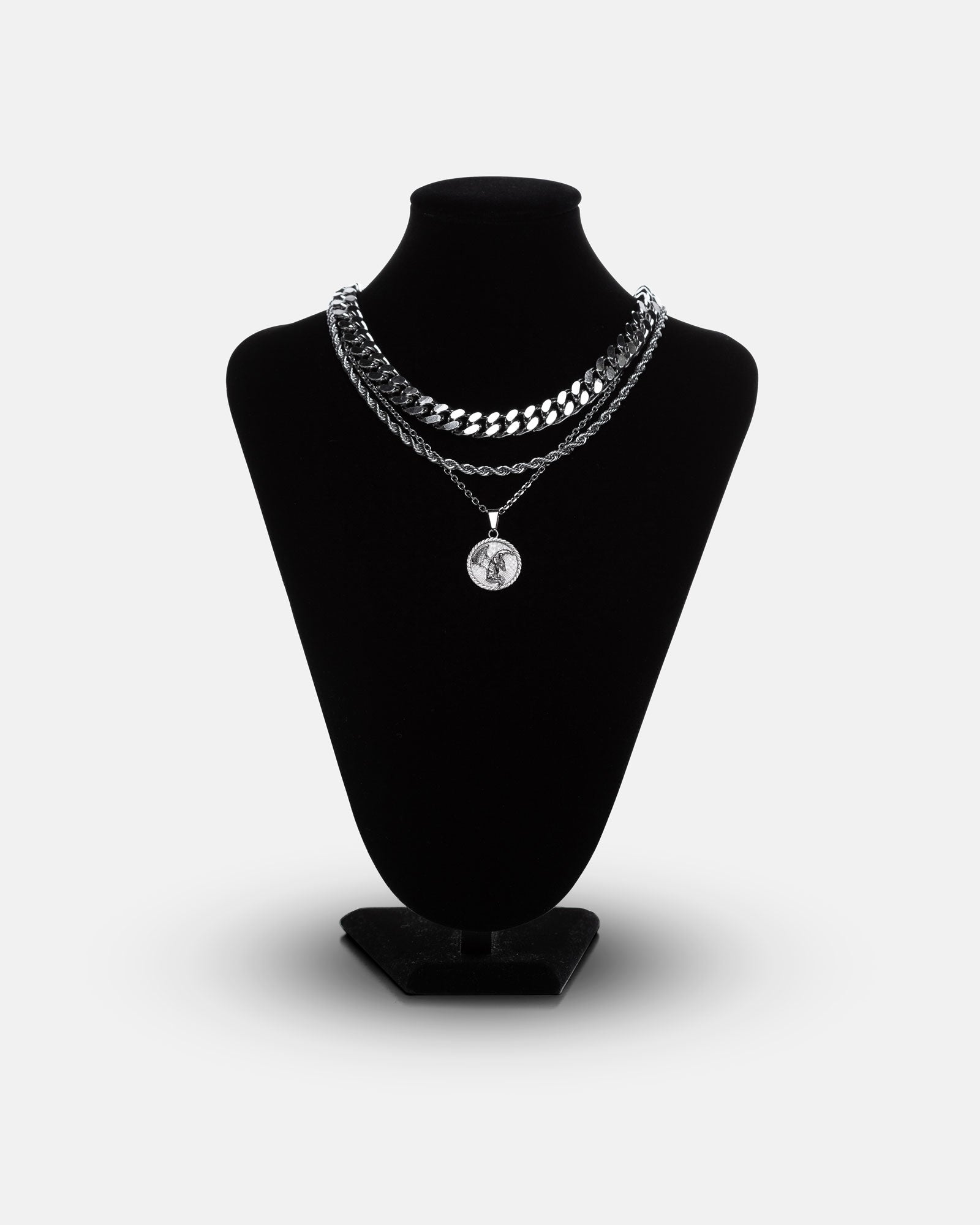 A black velvet necklace stand displays the Date Night Stack: three silver chains—two thick Cuban link necklaces and one medallion pendant with an engraved figure—against a plain white background.