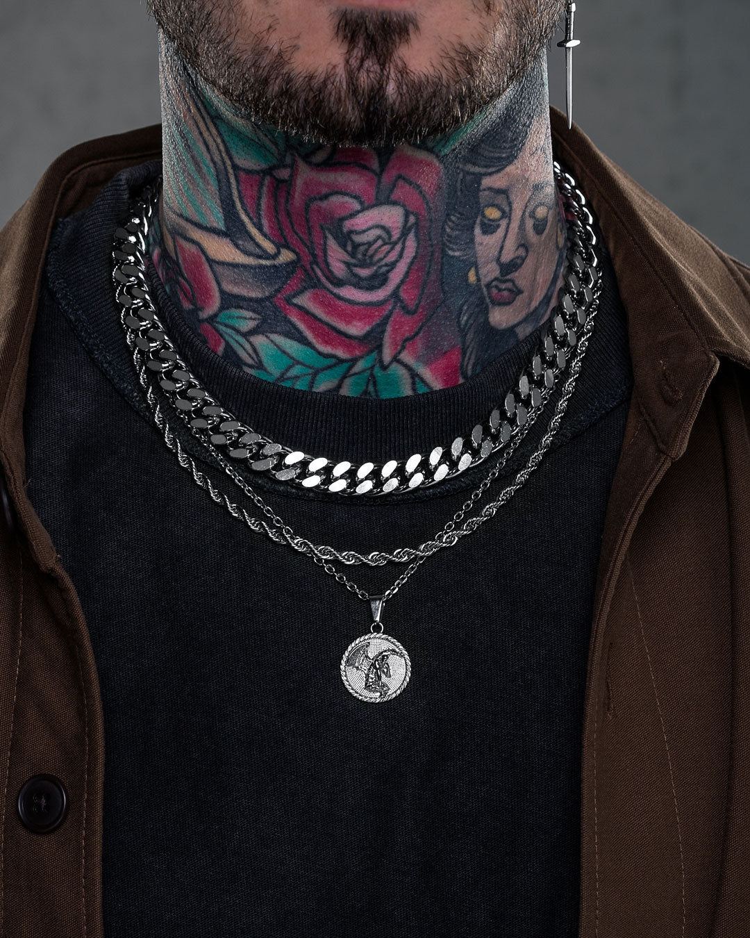 A close-up of a person wearing the Date Night Stack—layered silver chains with a Cuban link necklace and medallion pendant—over a black shirt, showcasing colorful tattoos and paired with a brown jacket.