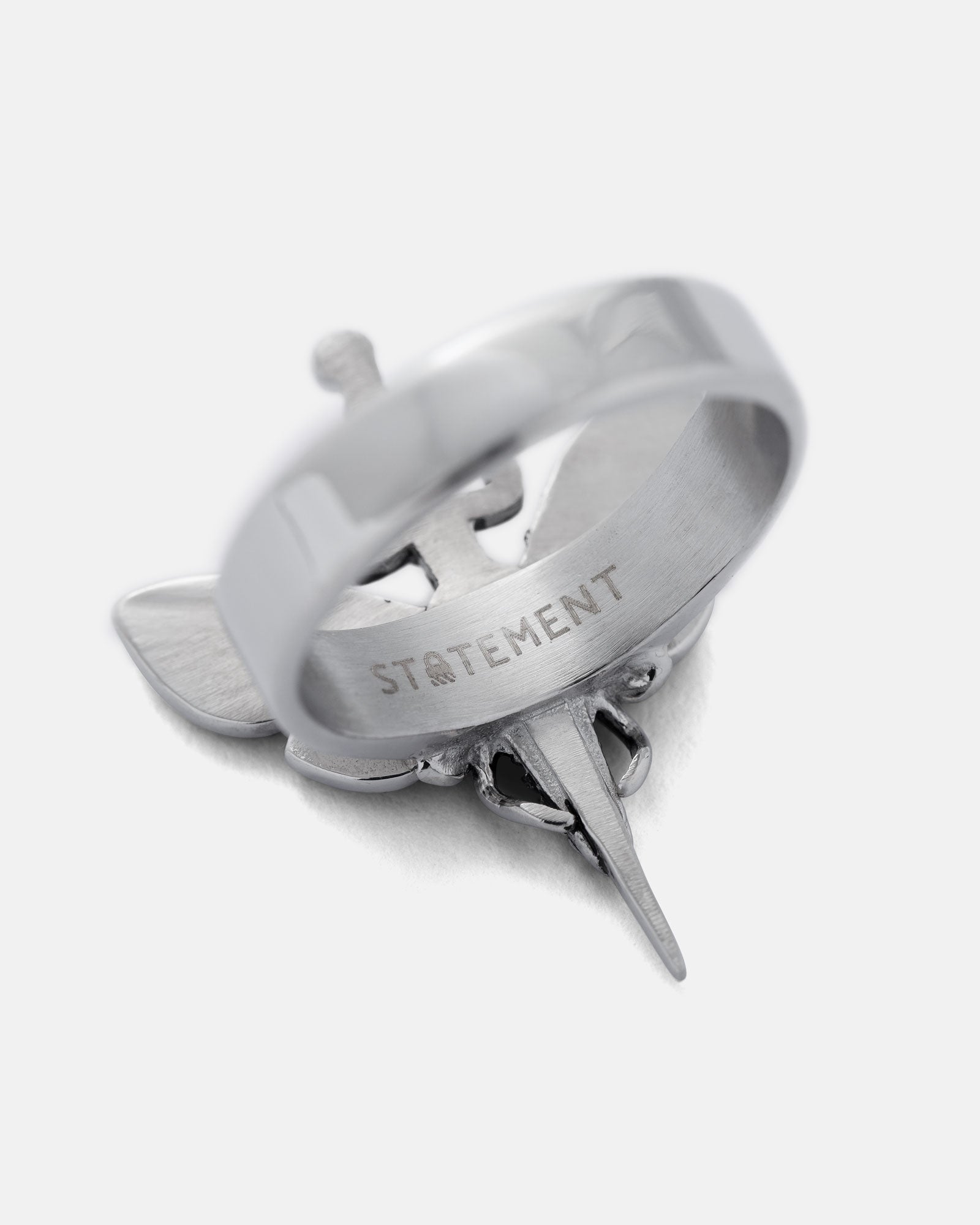 The Death's Head Moth Ring features a stainless steel band engraved with "STATEMENT," resting atop a sword-shaped pendant with wings, set against a plain white background.