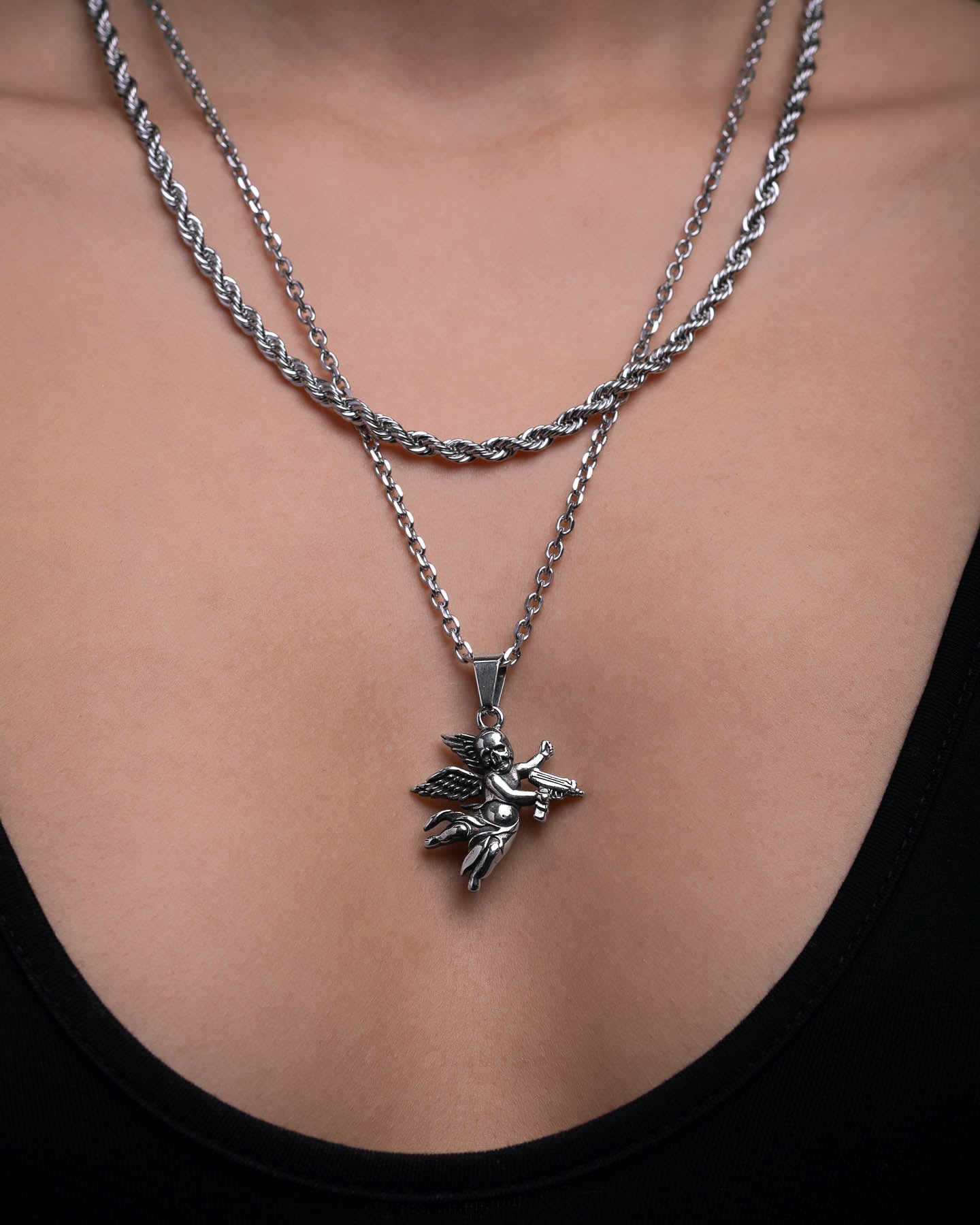A close-up of a person wearing two silver necklaces over bare skin: one twisted, and the other layered above a black deep-neckline top, featuring the Death's Cupid Pendant.