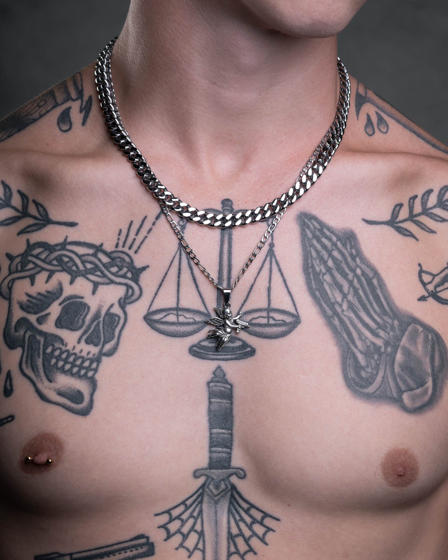 A tattooed chest displays a skull, praying hands, and scales. Two silver chains are worn, one featuring the Death's Cupid Pendant with a bold skull face. A nipple piercing is visible against the dark, neutral background.