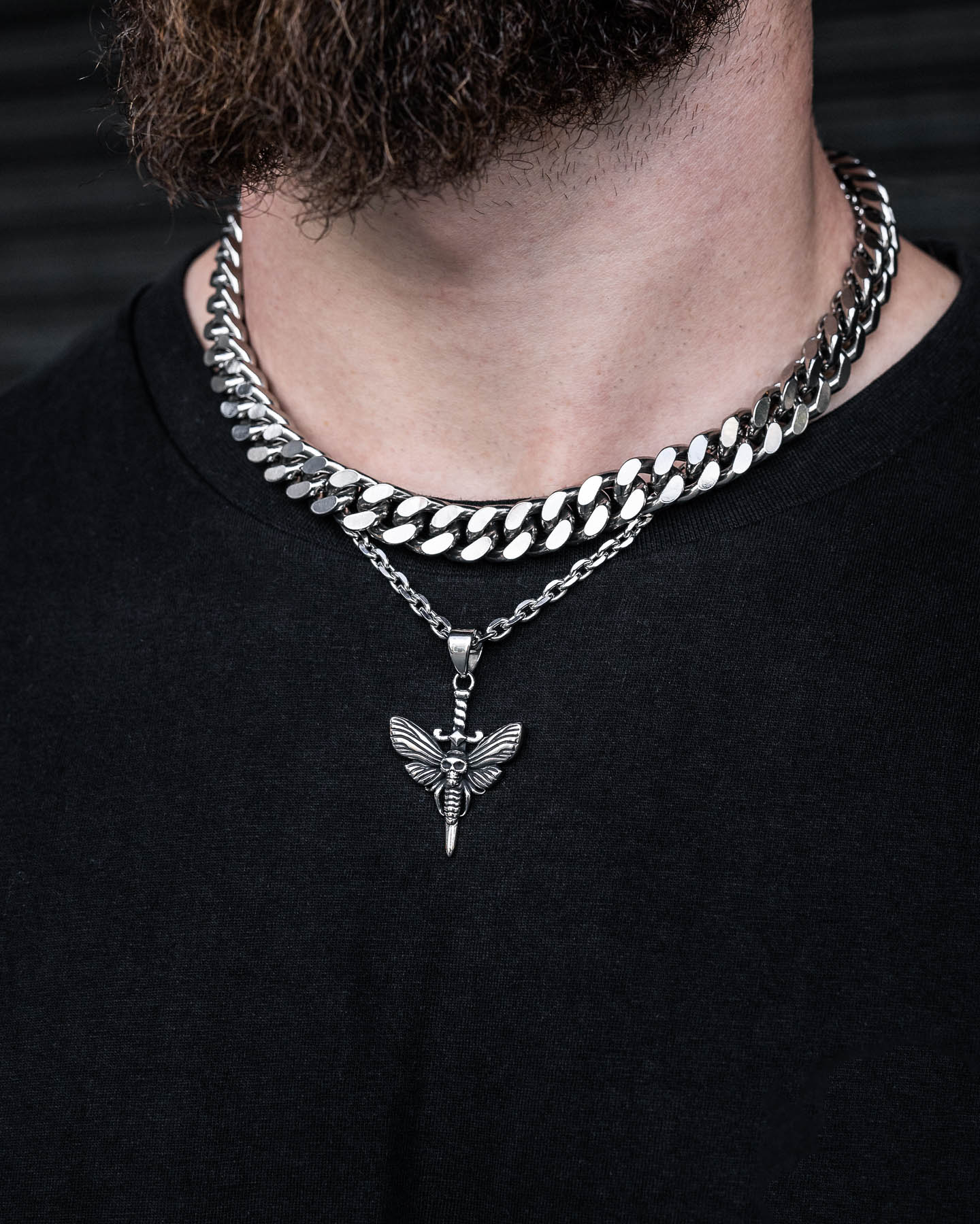 A person sports a thick silver chain and a slimmer necklace with the Death's Head Moth Pendant, inspired by Silence of the Lambs, over a black shirt. Only their lower face and beard are visible.