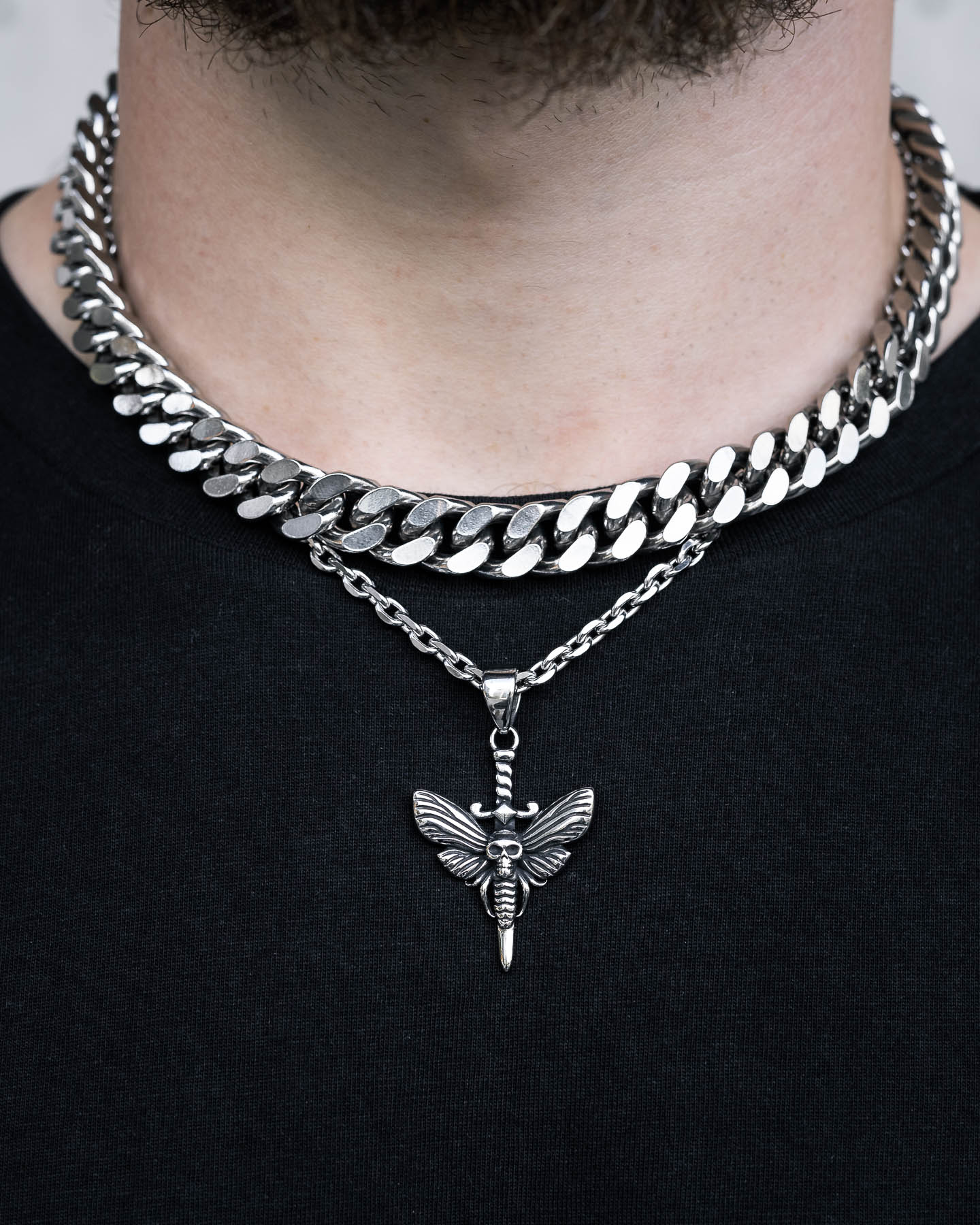 A person wears two silver necklaces—a thick chain and a thinner chain with the Death's Head Moth Pendant—over a black shirt. Only their neck and part of their chin are visible.