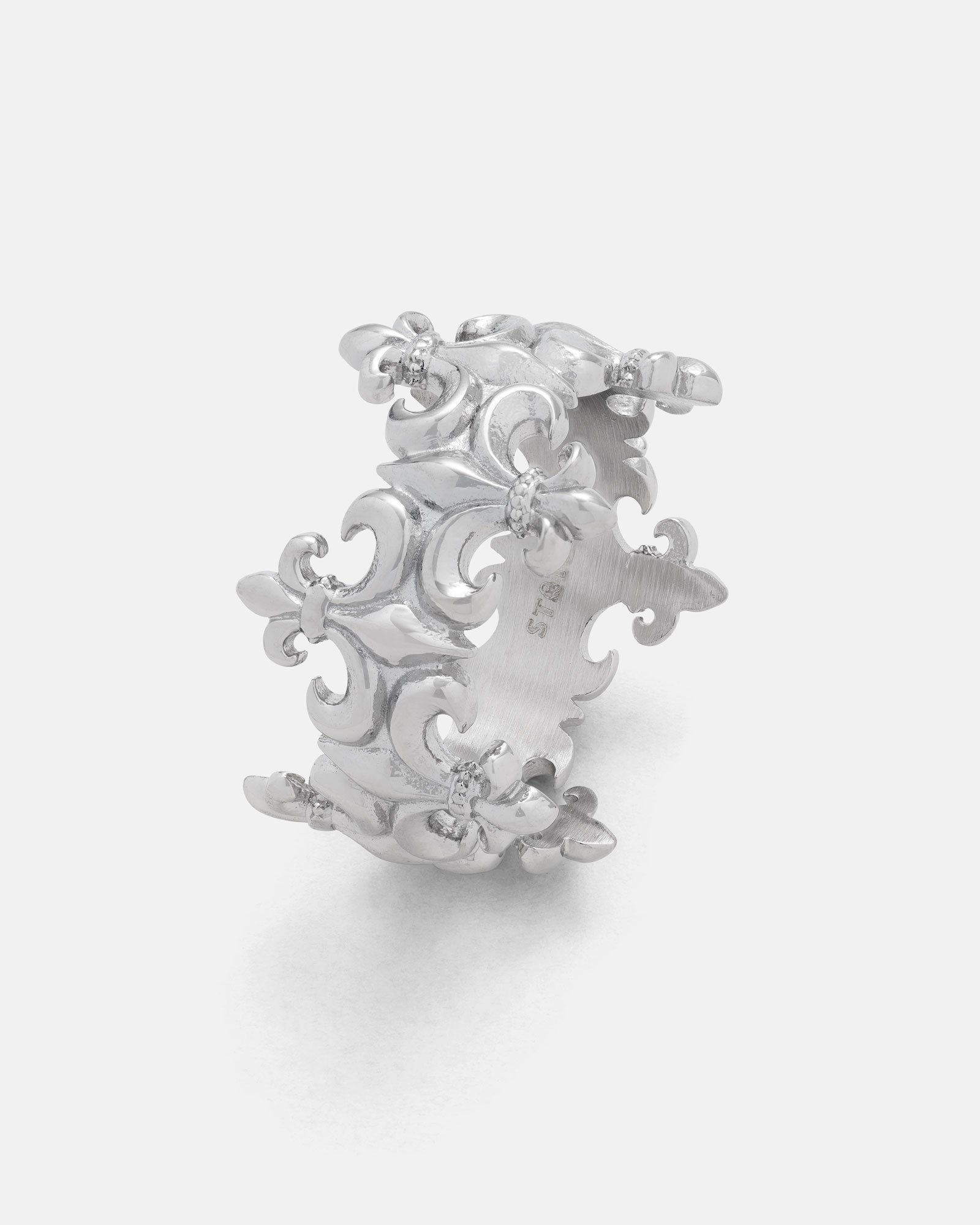 Introducing the “Crested” Fleur De Lis Ring by Statement Collective—a striking crown-like band crafted from durable 316L stainless steel. This silver ring showcases an intricate, repeating fleur-de-lis pattern, with small round gemstones delicately set at the intersections, resulting in an ornate and elegant design. Perfectly styled against a plain, light-colored background, it makes a memorable statement piece.