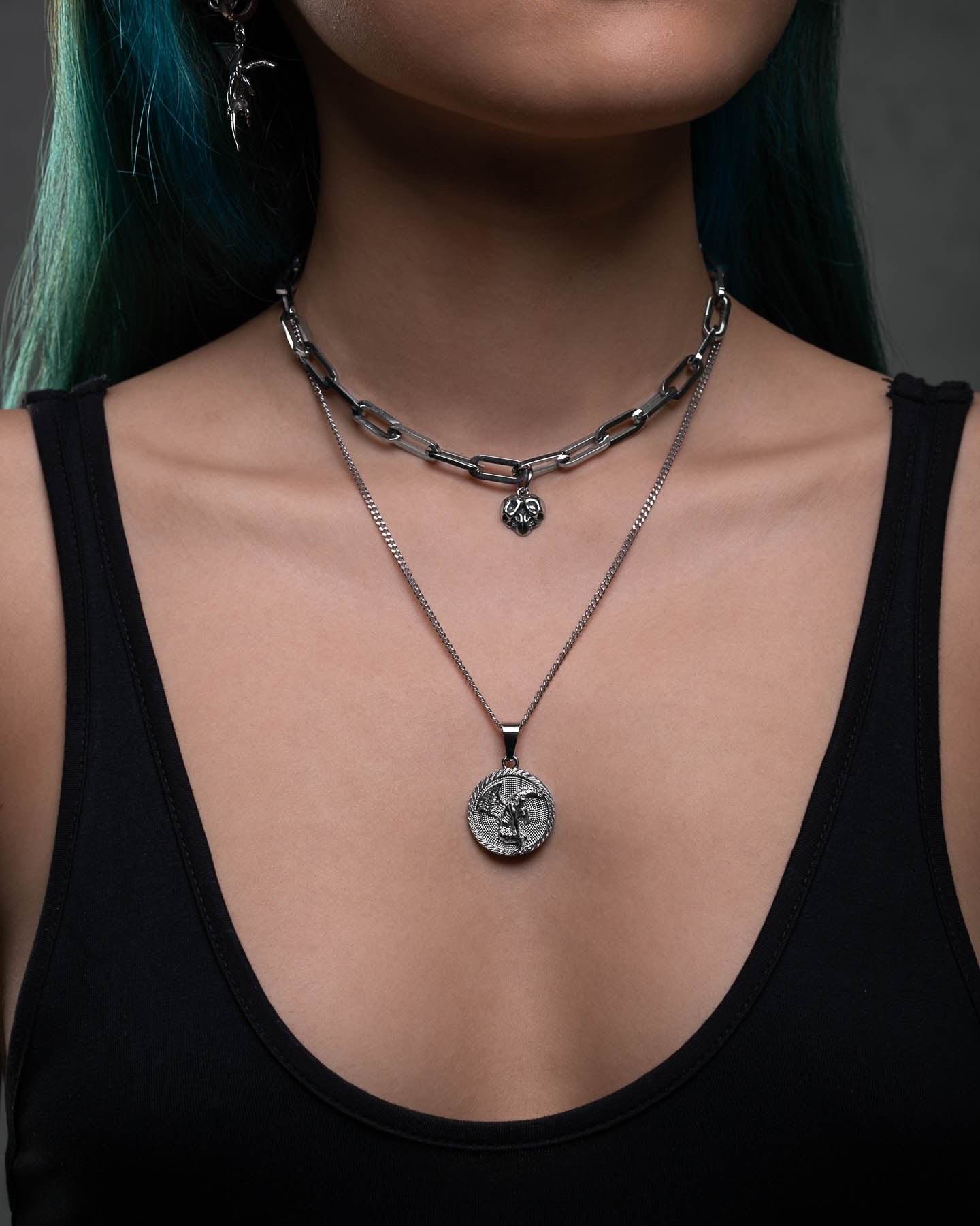 A person with blue-green hair wears a black tank top and layered silver necklaces, including a chunky chain, small pendant, and the Fallen Angel Medallion Pendant featuring an engraved figure.