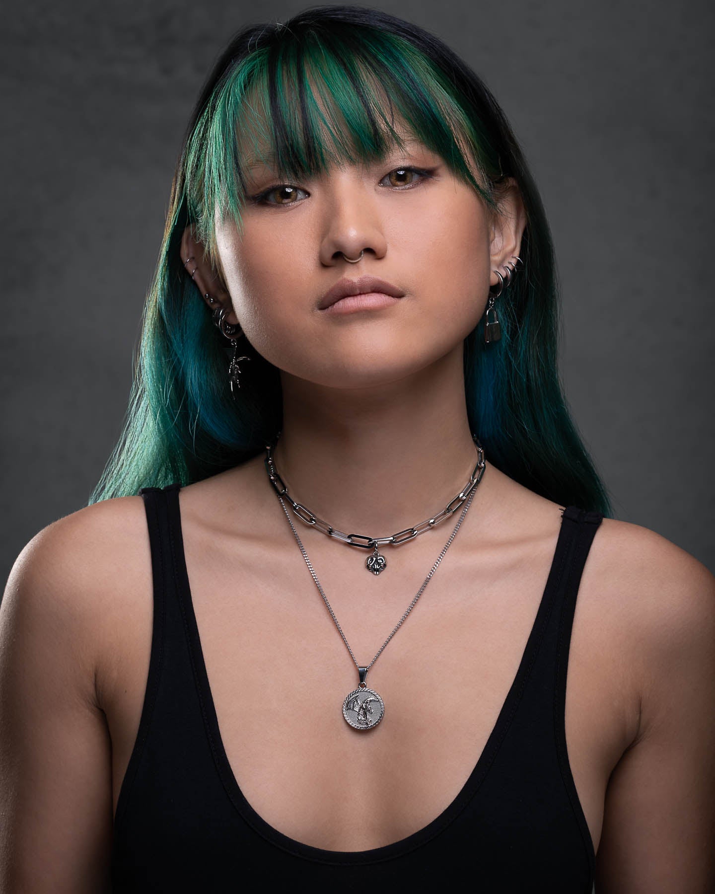 A young woman with long green hair and bangs stares confidently at the camera. She wears a black tank top, layered silver necklaces including the Fallen Angel Medallion Pendant, and multiple earrings. The background is dark and plain.
