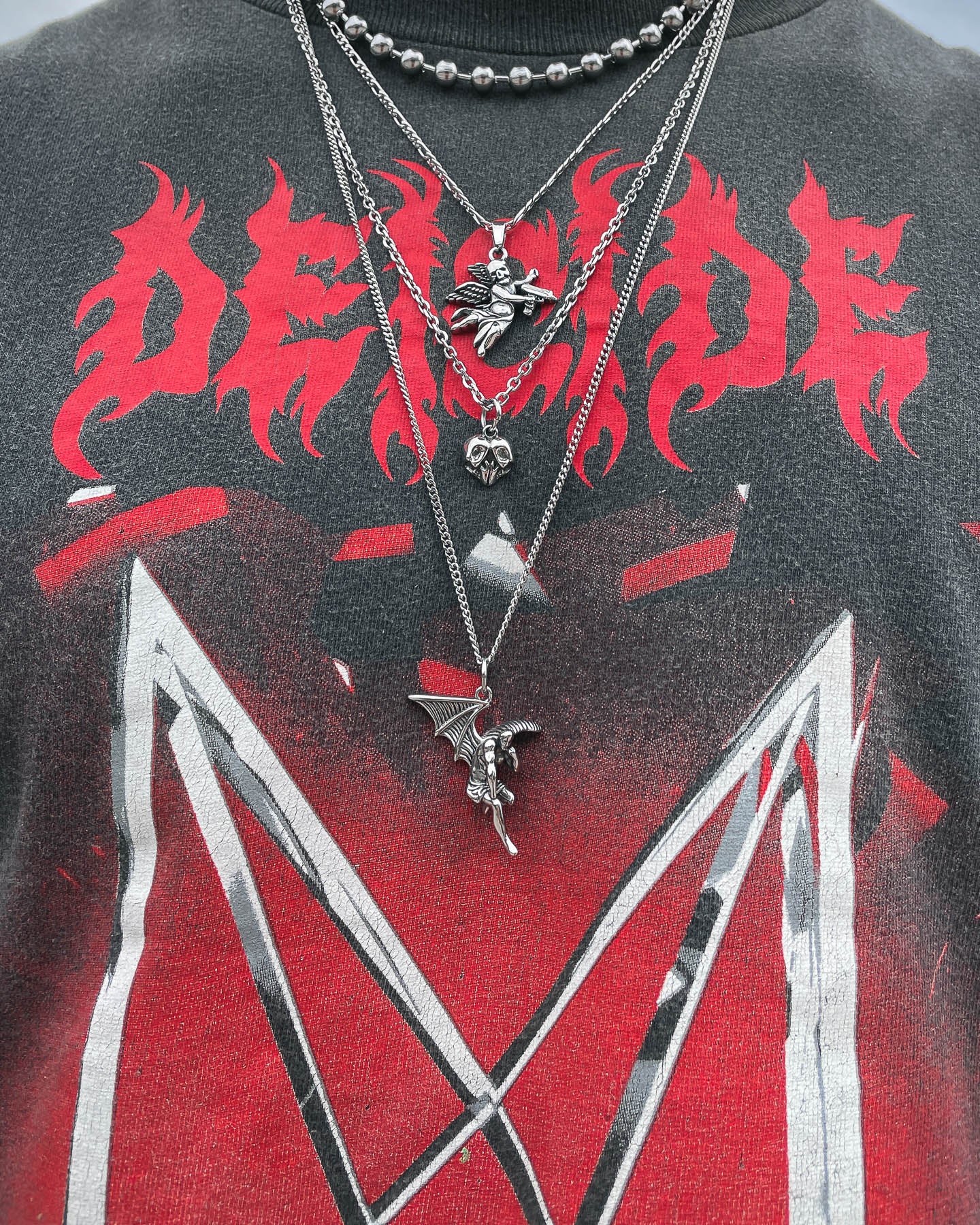 A close-up of a black shirt with red and white graphic designs reading DEICIDE in fiery red, accessorized with layered silver jewelry featuring the Fallen Angel Pendant along with skull, dragon, and heart necklaces.