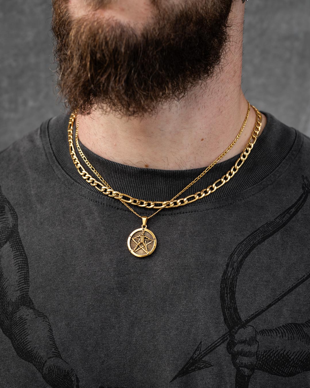 A bearded person in a dark shirt with a faint archer graphic wears two gold chains, including the Statement Collective Figaro Set (Custom Pendant Choice) in gold with an 18k pendant featuring a star design.