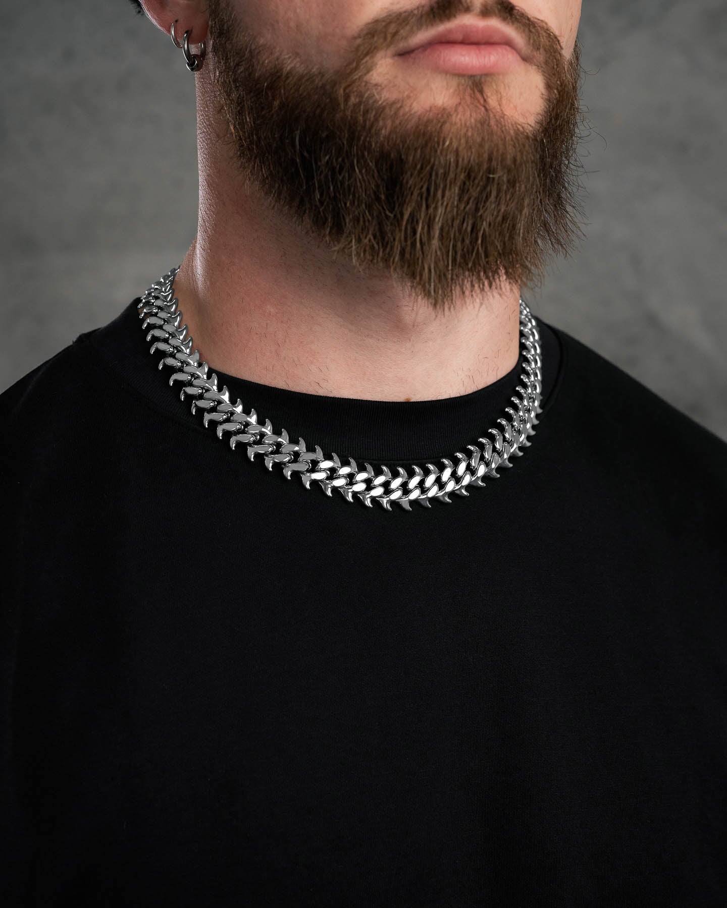 A man with a thick brown beard wears a "Fishbone" Curved Spike Cuban Chain and a plain black shirt, set against a gray background.
