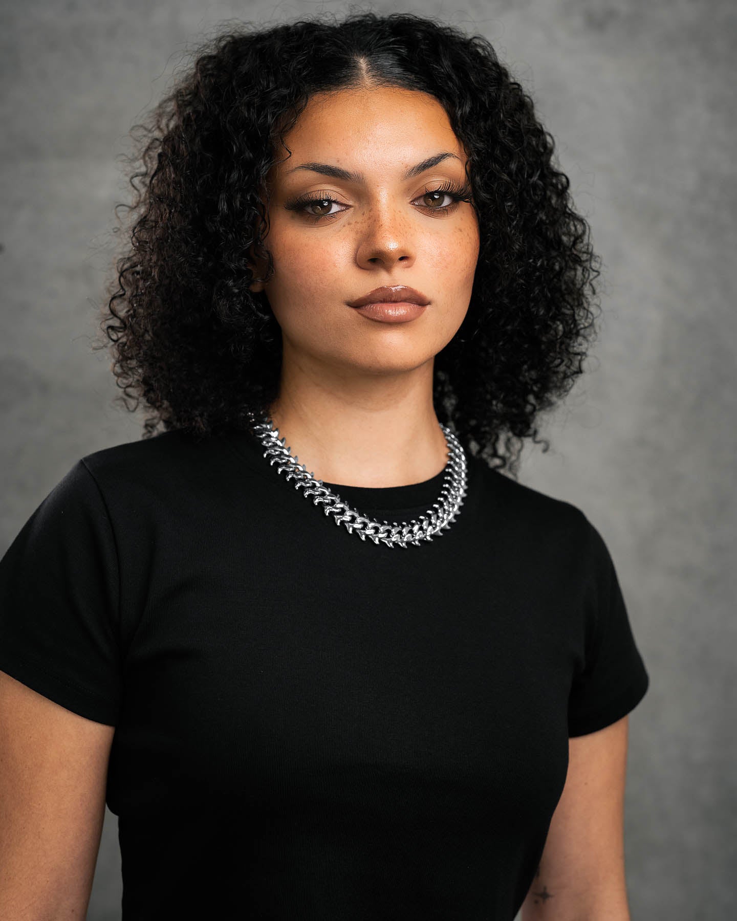 A woman with curly dark hair wears a black t-shirt and the "Fishbone" Curved Spike Cuban Chain, standing confidently against a gray textured background.