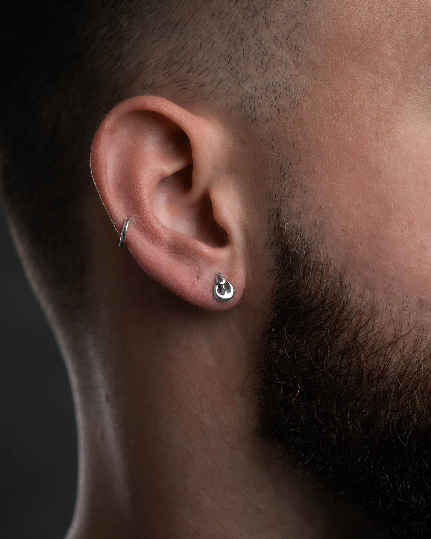 Close-up of a person's head showing two stainless steel earrings—a small hoop in the upper ear and a hypoallergenic Flame Stud Earring in the earlobe. The person has short hair and a trimmed beard.