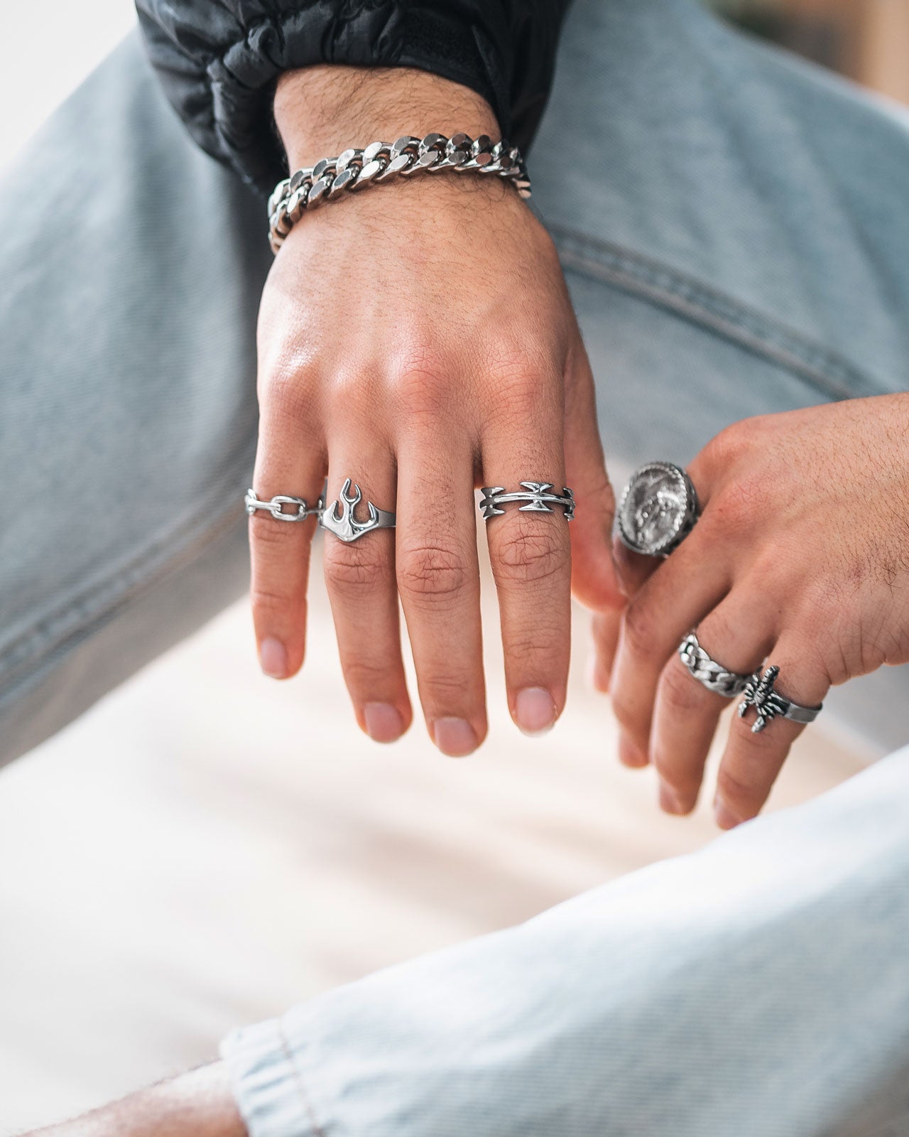 A close-up of two hands showcases several silver rings, including the Flame Ring, and chunky streetwear accessories; the person is dressed in light blue jeans and a black jacket.