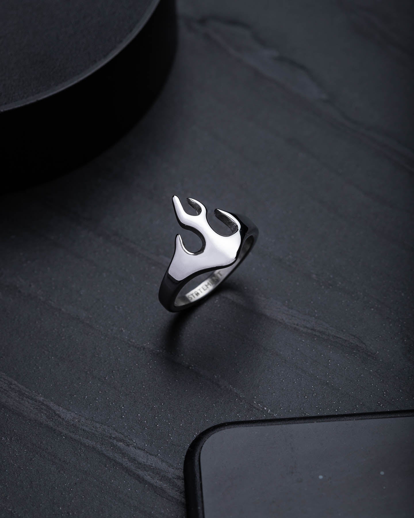 The Flame Ring, featuring a polished silver finish and upright flame-shaped design, is displayed on a dark textured surface—ideal for adding bold streetwear style.