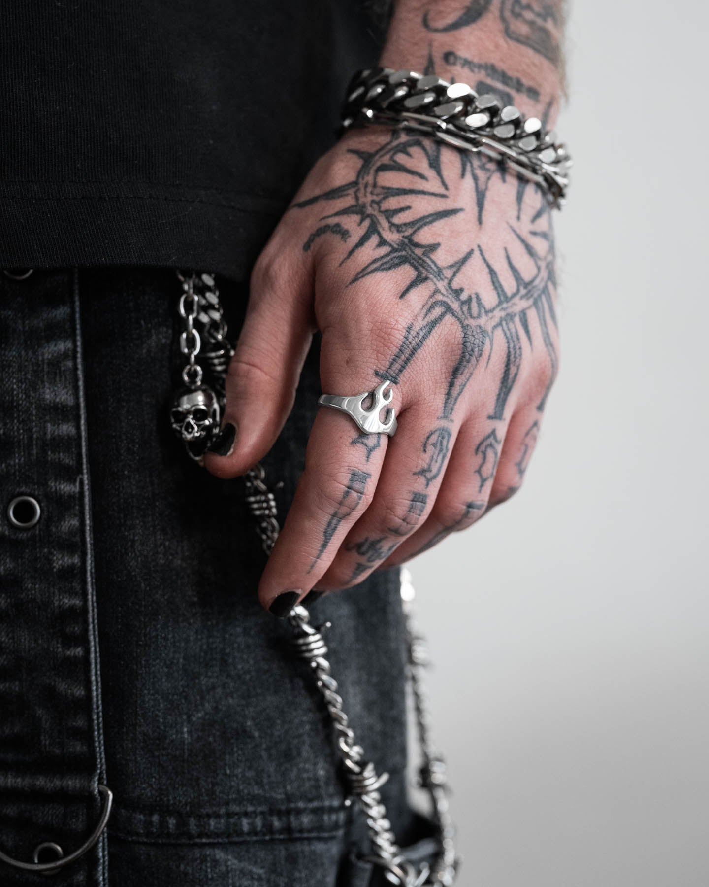 A tattooed hand with black nail polish wears a chunky silver bracelet, chain, and the Flame Ring. Dressed in black denim with metal details, a skull chain streetwear accessory hangs from their belt.