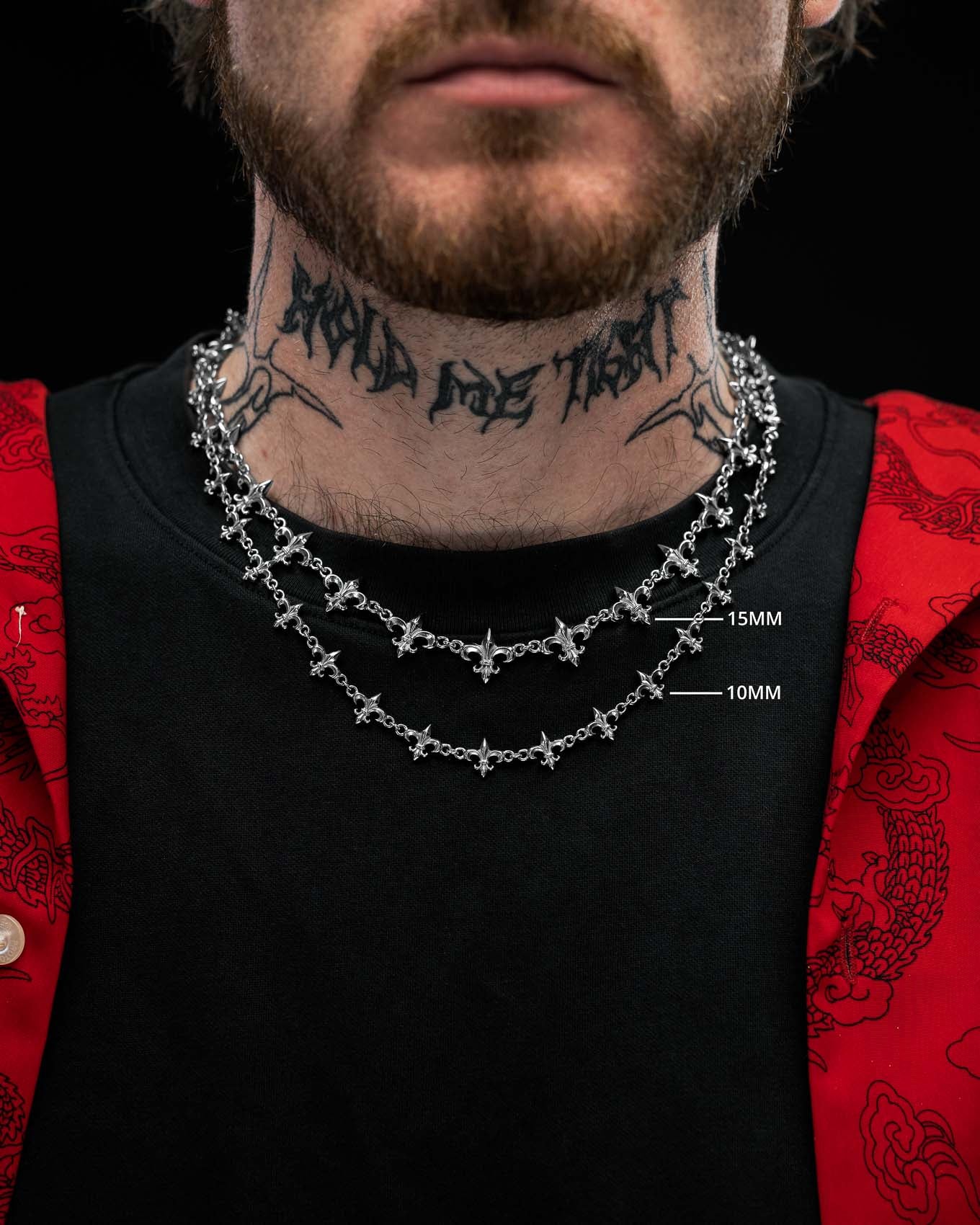 Close-up of a person wearing two silver cross necklaces of different thicknesses, including the “Fleur De Lis” 10mm Chain by Statement Collective, with a tattooed neck, black shirt, and a red jacket featuring dragon patterns.