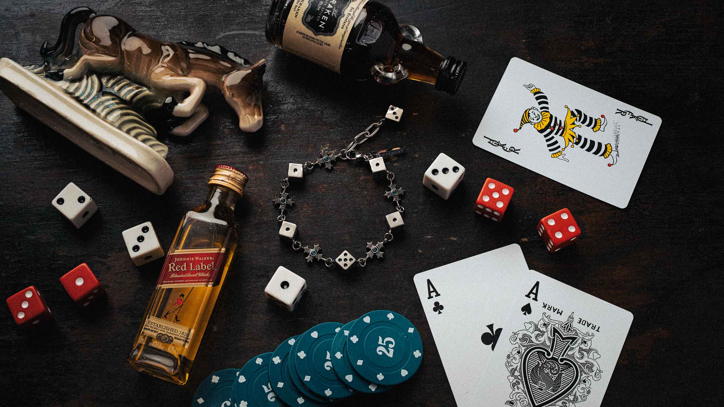 A styled flat-lay of poker and drinking props on a dark wooden surface, including two bottles, playing cards, dice, chips, a bracelet, and a horse figurine.
